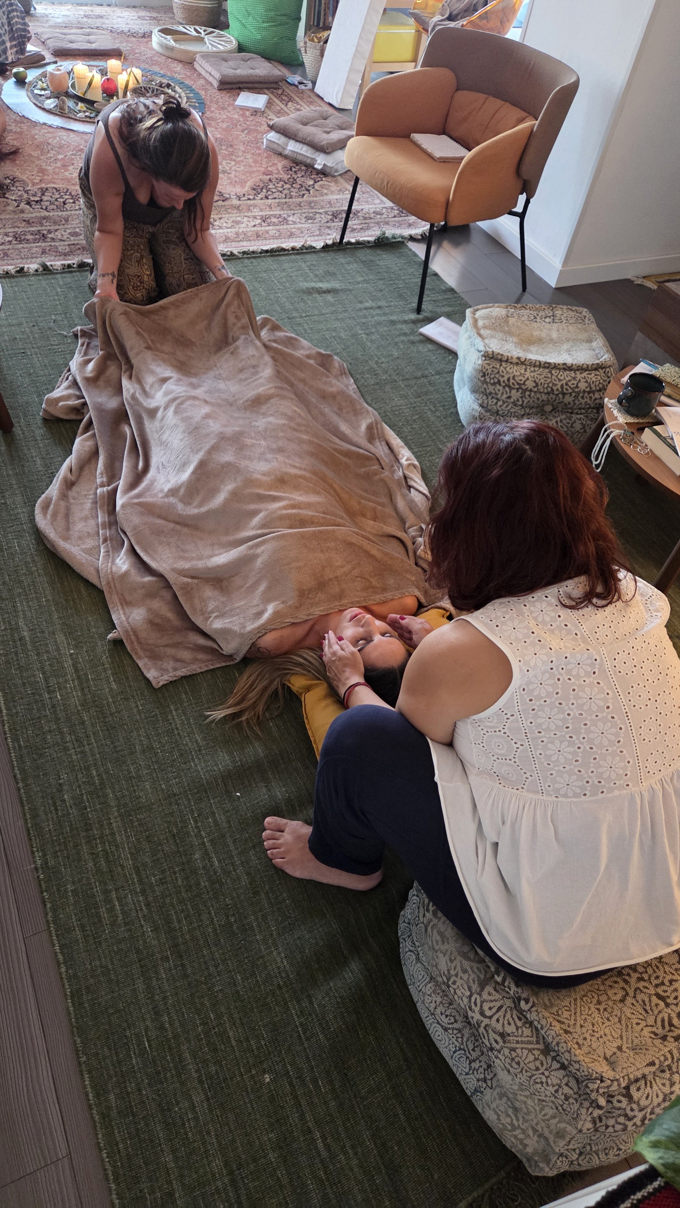 A person lying on the floor covered with a blanket, receiving support from another person kneeling beside them, with a third person attending to their head, in a cozy room with cushions, chairs, and a decorative rug.