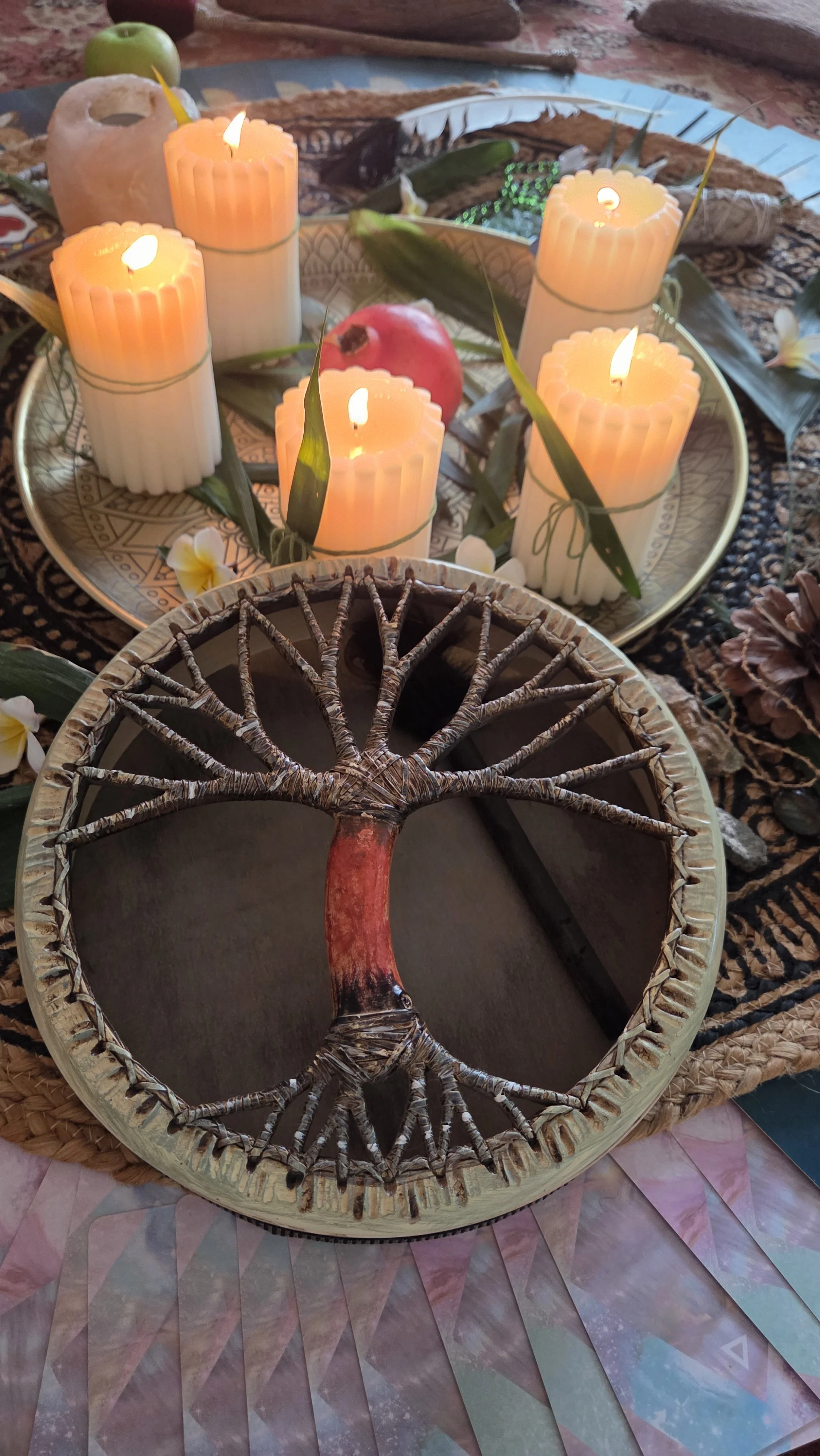 A decorated tray with lit candles, green leaves, an apple, and flowers, with a traditional drum in the foreground and a patterned cloth underneath.