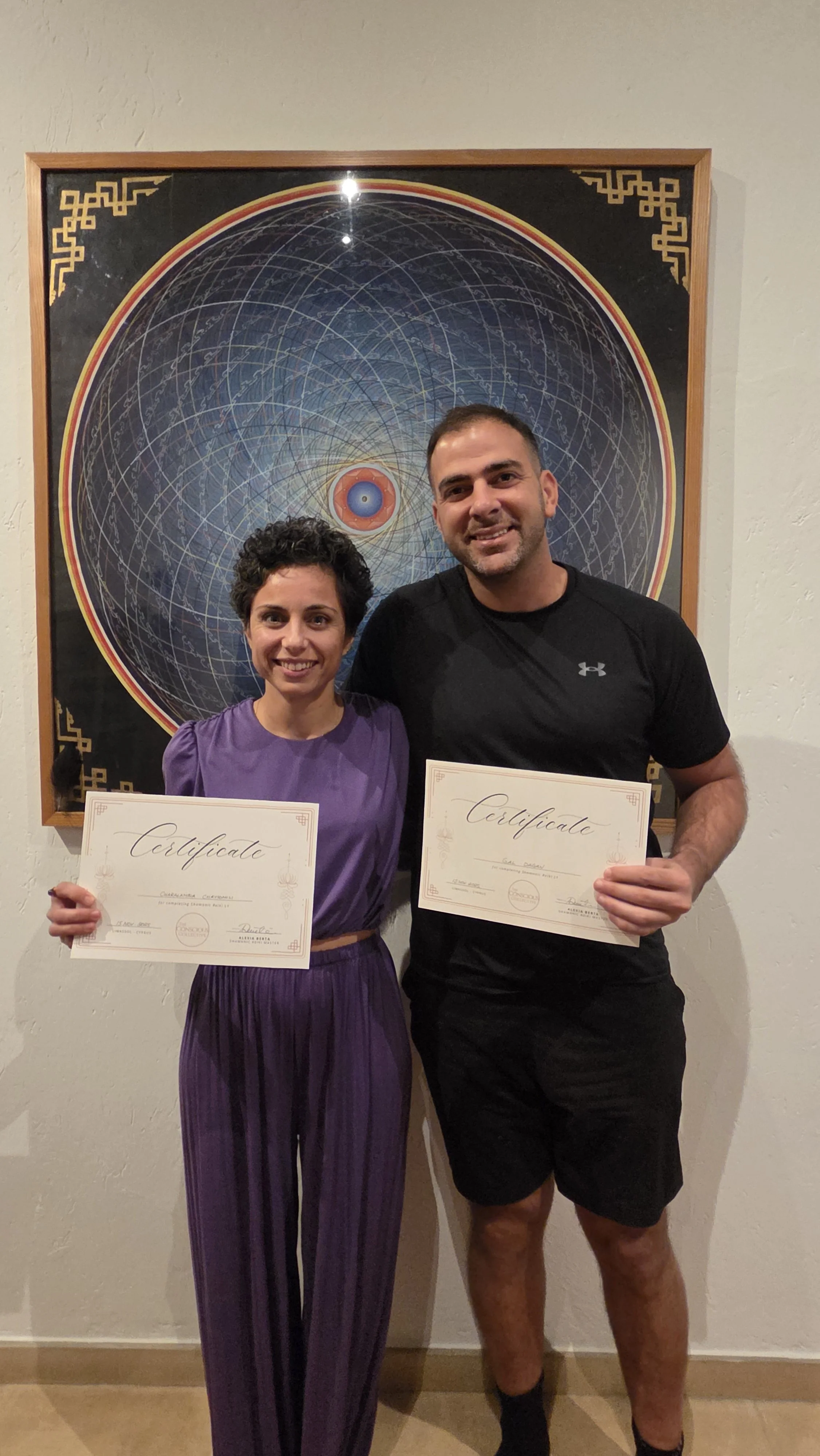 A woman and a man standing in front of a large artistic poster at an indoor event, holding certificates and smiling at the camera.