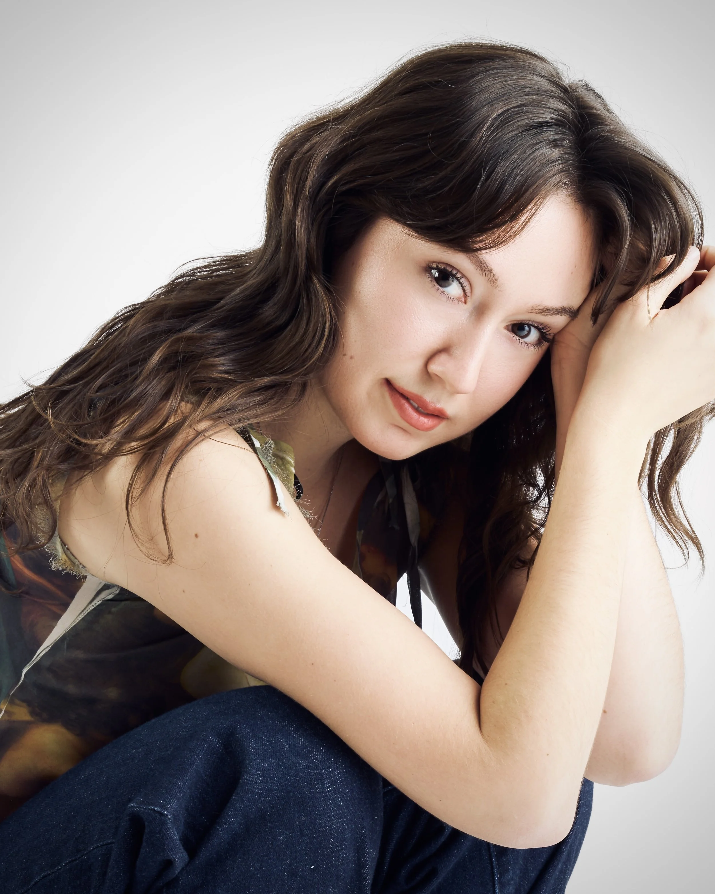 Close-up of a woman with wavy brown hair, light skin, and blue eyes, smiling softly with one hand resting near her head, wearing casual clothing against a plain background.
