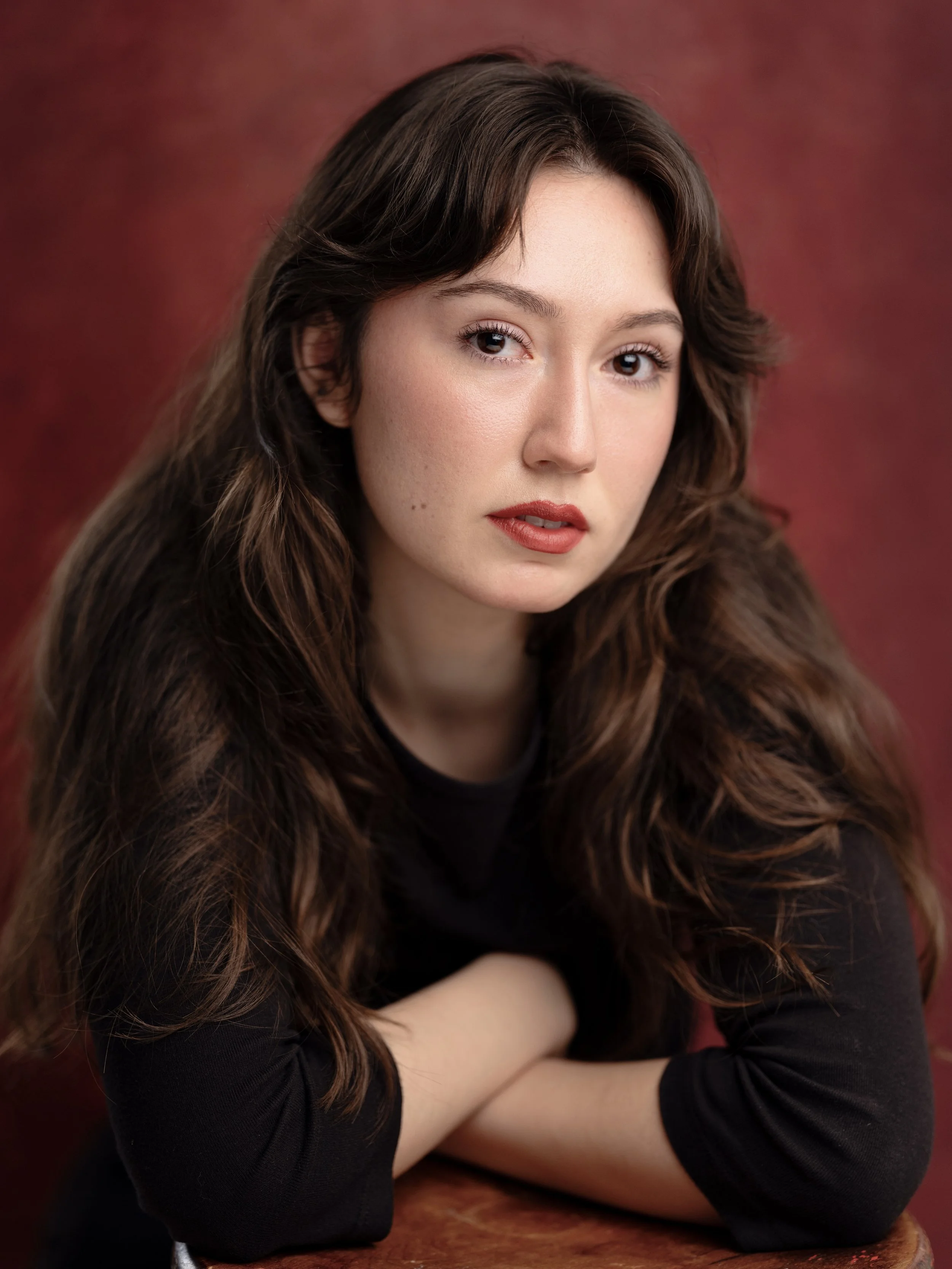 Close-up of a young woman with long wavy brown hair, light skin, and red lipstick, resting her arms on a wooden surface, against a blurred red background.