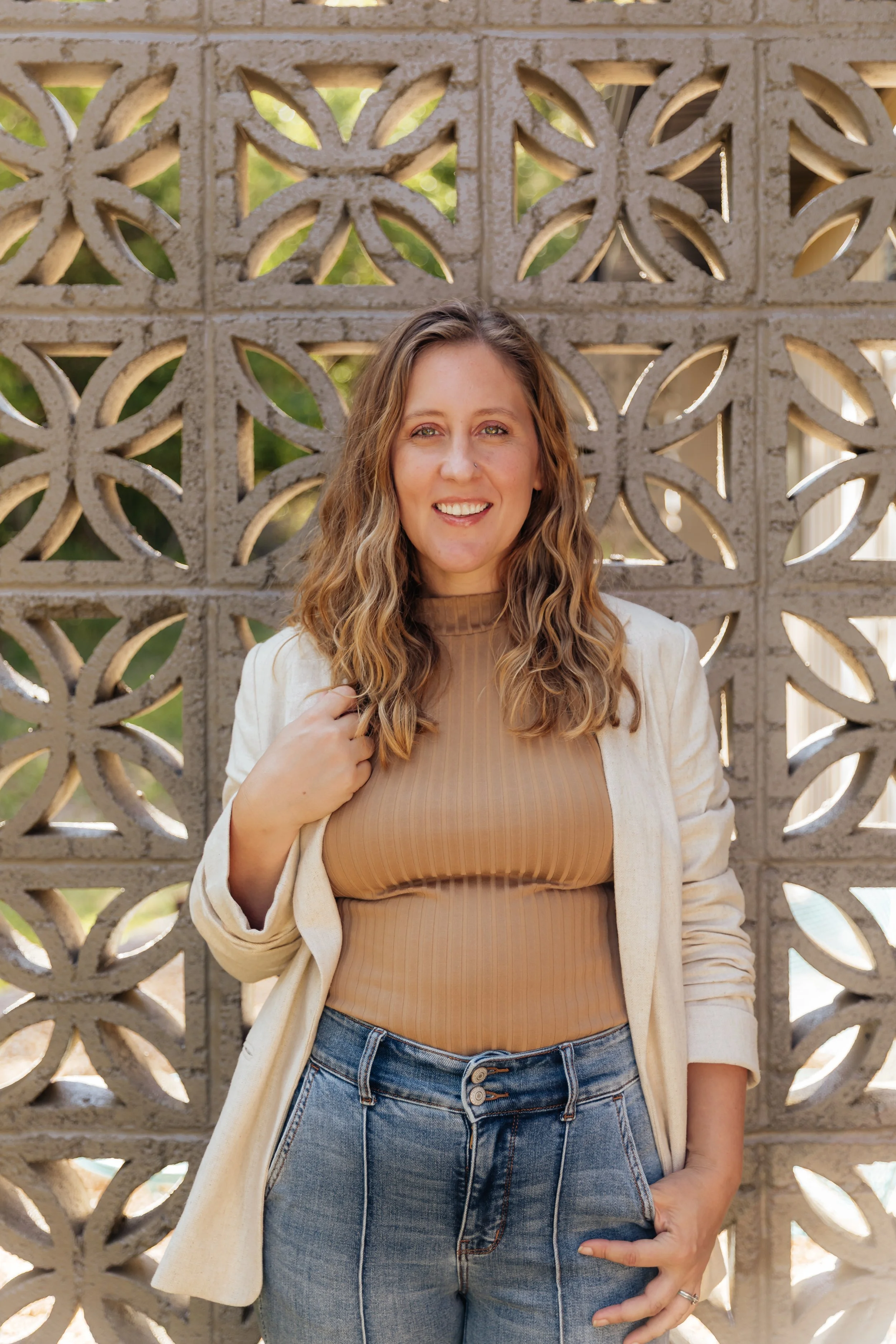 A woman with wavy, shoulder-length hair smiling, standing in front of a decorative concrete block wall, wearing a beige blazer over a tan ribbed turtleneck and blue jeans.