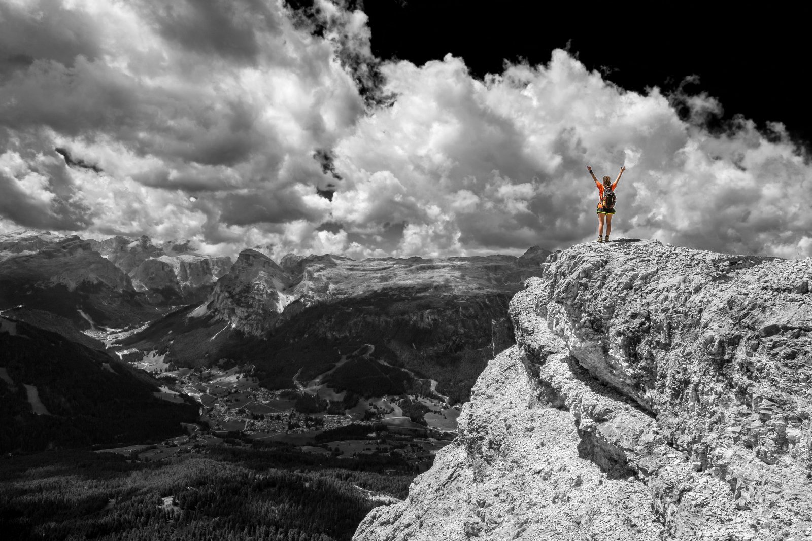 Person standing on a rocky mountain summit with arms raised, overlooking a valley and mountains under cloudy sky.