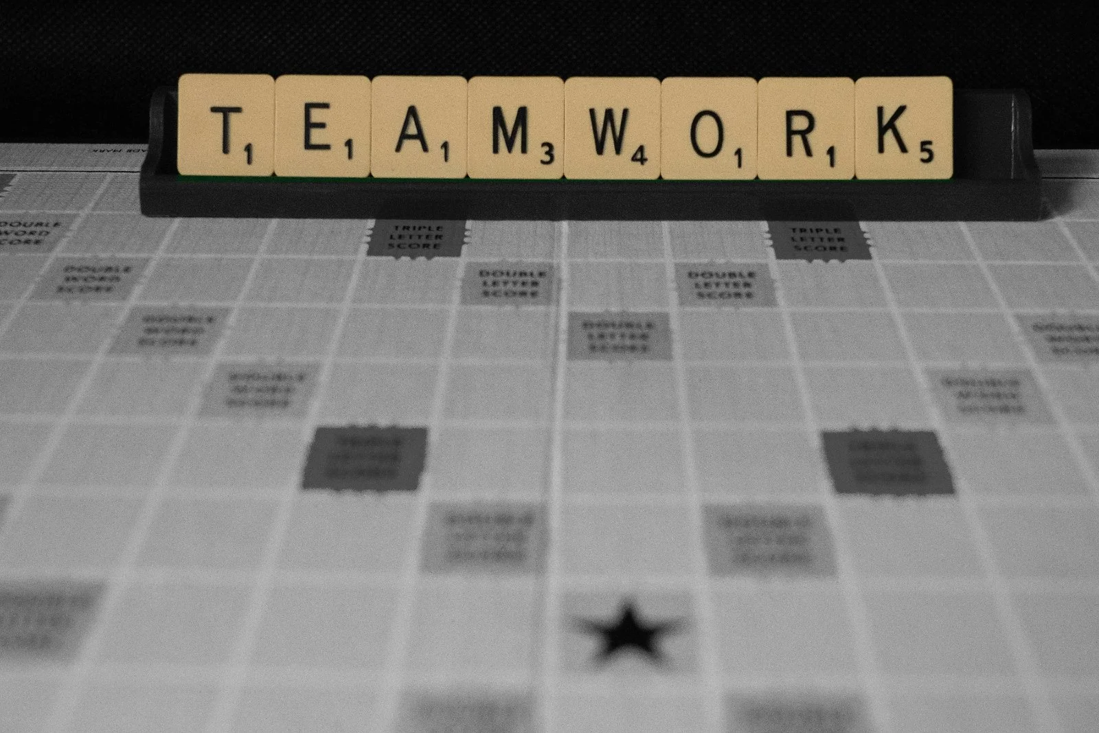Scrabble tiles arranged to spell 'TEAMWORK' on a black tile rack, placed on a game board with visible grid and scoring squares.