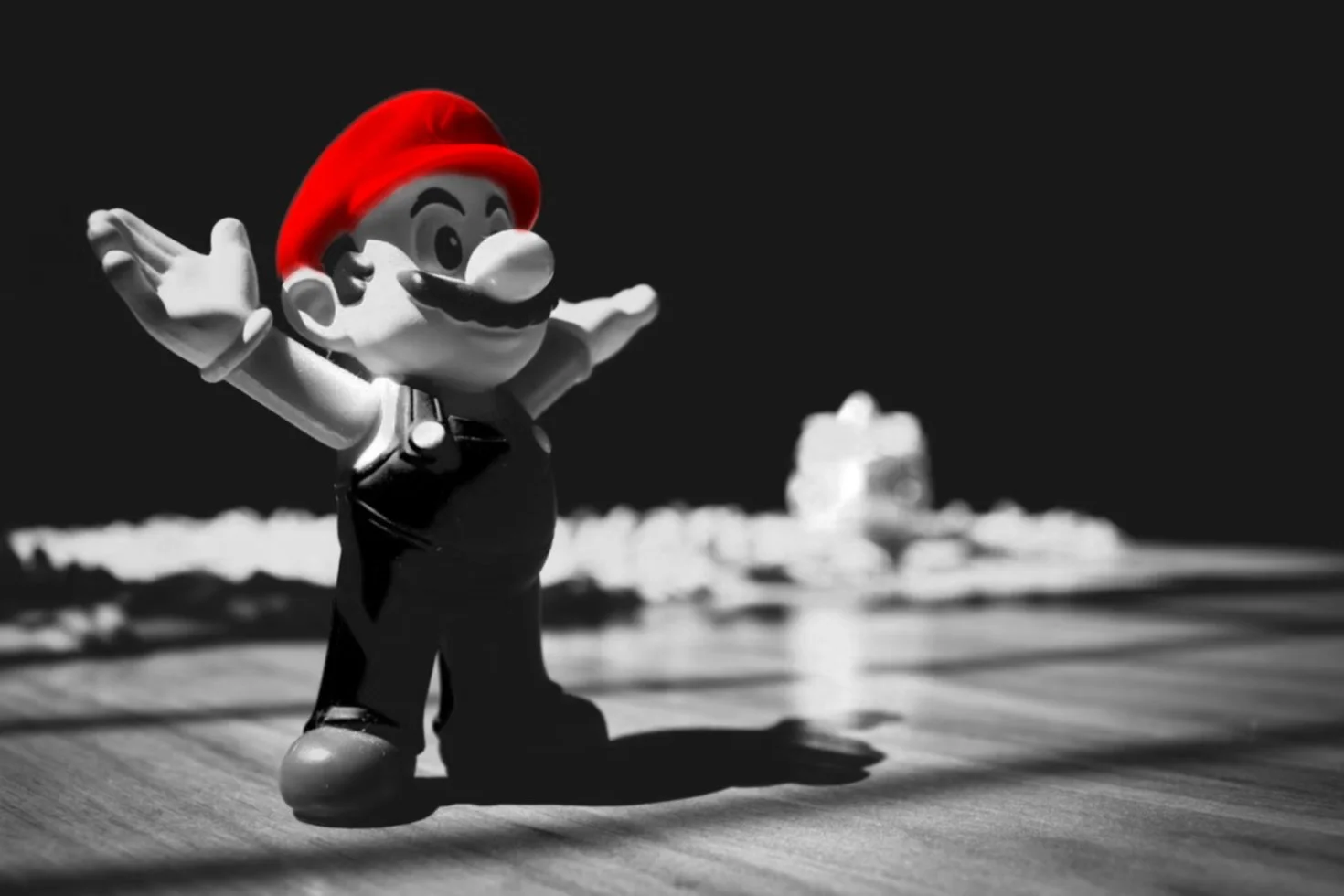 A toy figure of Mario with his arms outstretched in front of a black and white background, with a blurred object in the distance.
