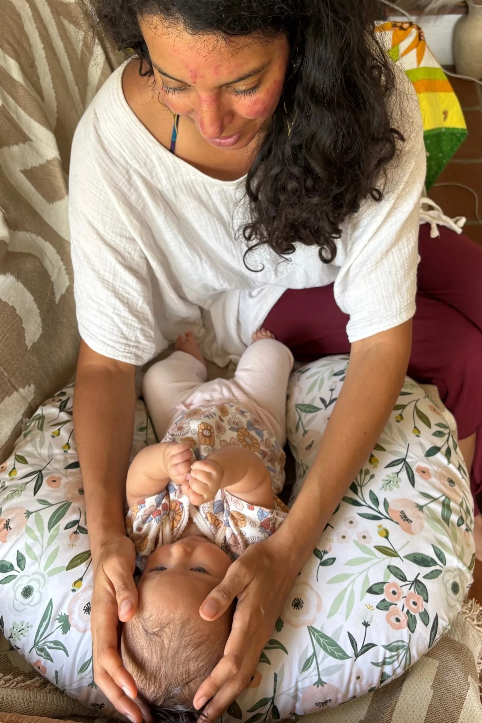 A woman with long dark curly hair and a toddler with blonde hair are sitting on a floral sheet, with the woman gently holding the child's head. The woman has some acne scars on her face and is wearing a white shirt. The toddler is looking up while holding their hands together near their chin, wearing a light-colored shirt with a floral pattern. They are in a cozy indoor setting.