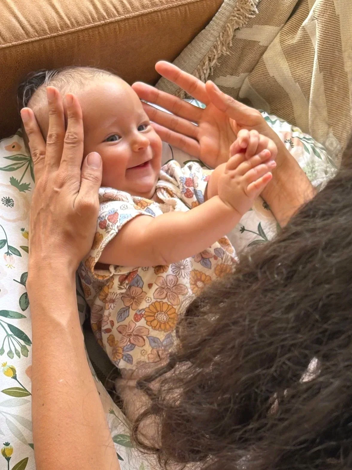 A smiling baby with blue eyes lying on a floral pillow, holding hands with an adult, while the adult gently touches the baby's head and face in a cozy indoor setting.