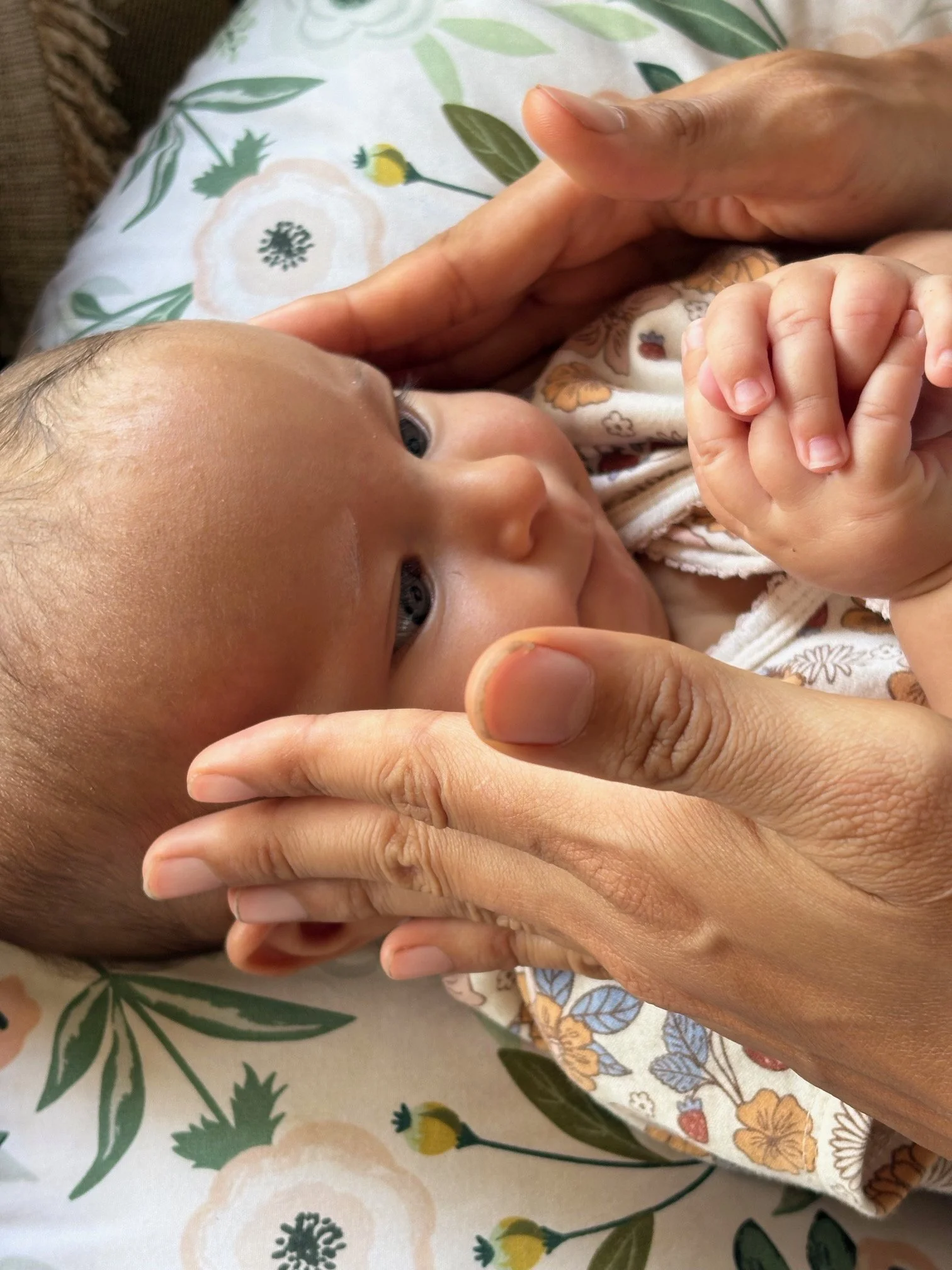 A close-up of an adult holding a baby's hand and a finger. The baby is lying on a floral-patterned surface with leaves and flowers. The adult's hand gently supports the baby's head and hand.