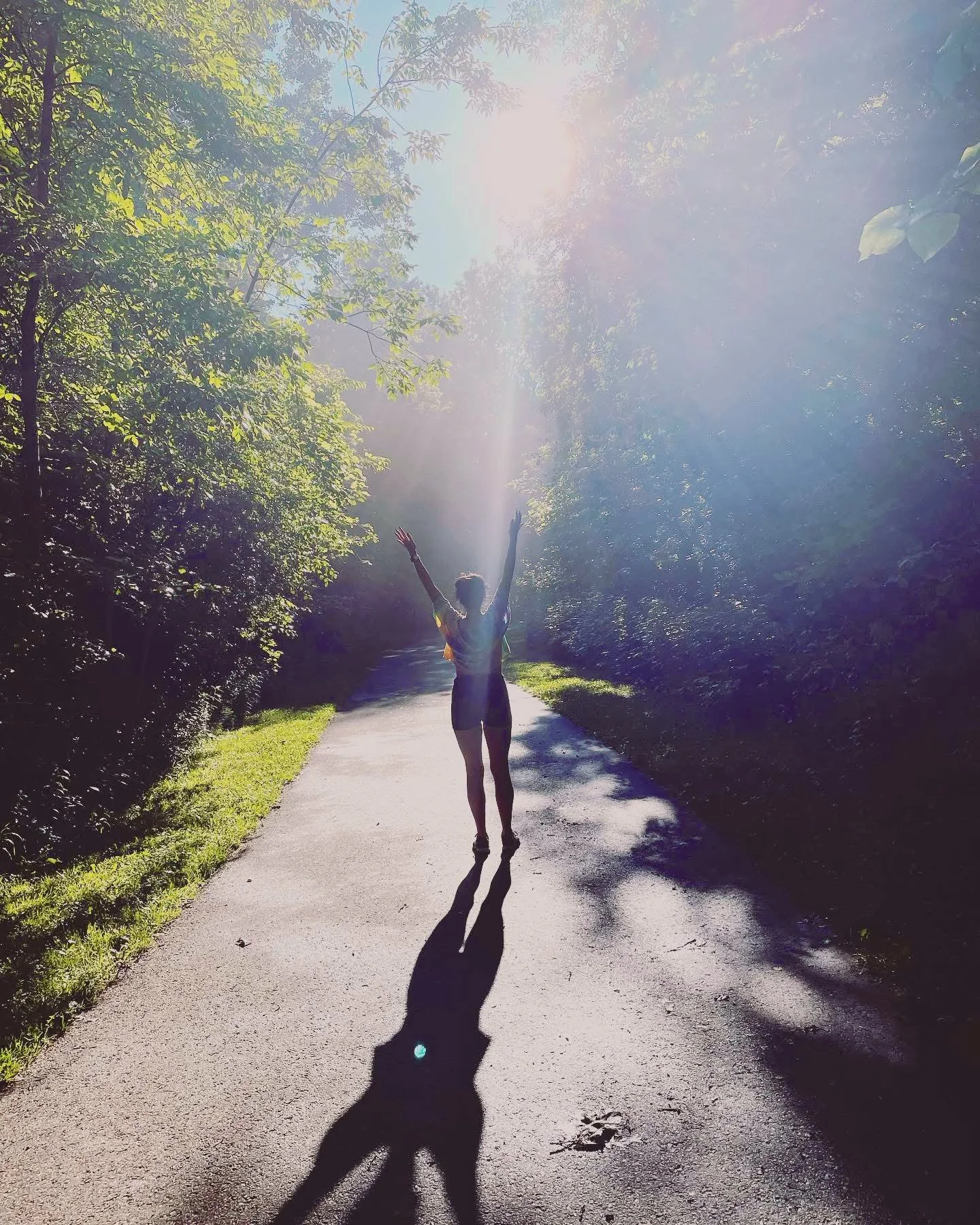 A person standing on a sunlit forest path with arms raised, casting a shadow on the ground, surrounded by green trees and bright sunlight filtering through the leaves.