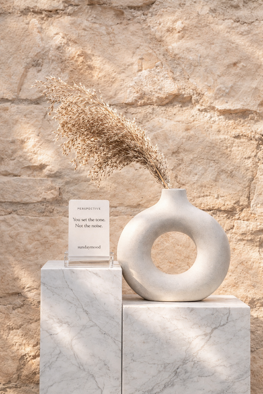 A white ceramic vase with a large circular hole in the center, holding dried beige pampas grass, is placed on a white marble pedestal against a textured beige stone wall. A small sign next to the vase reads: "PERSPECTIVE: You set the tone. Not the noise. sundaymood."