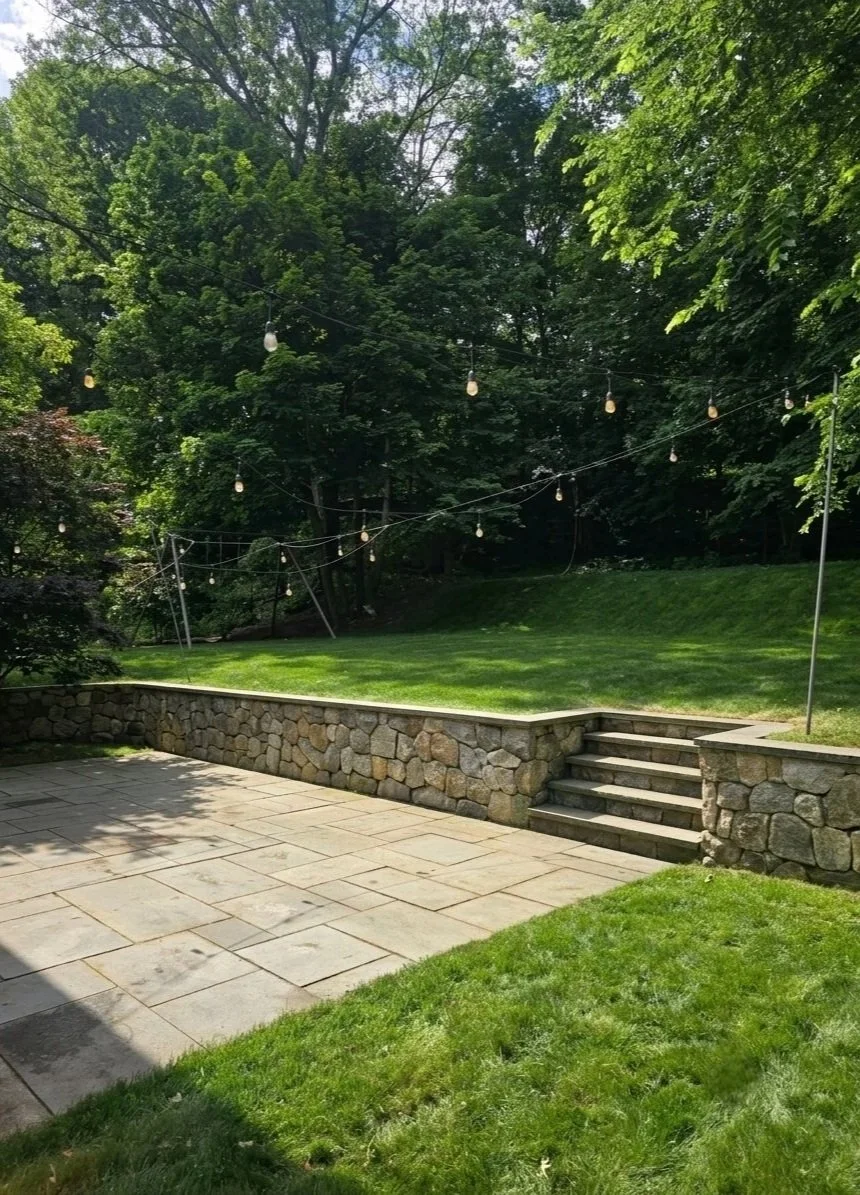 A back yard with a bluestone patio, a stone retaining wall and a green lawn in Brookline, MA.