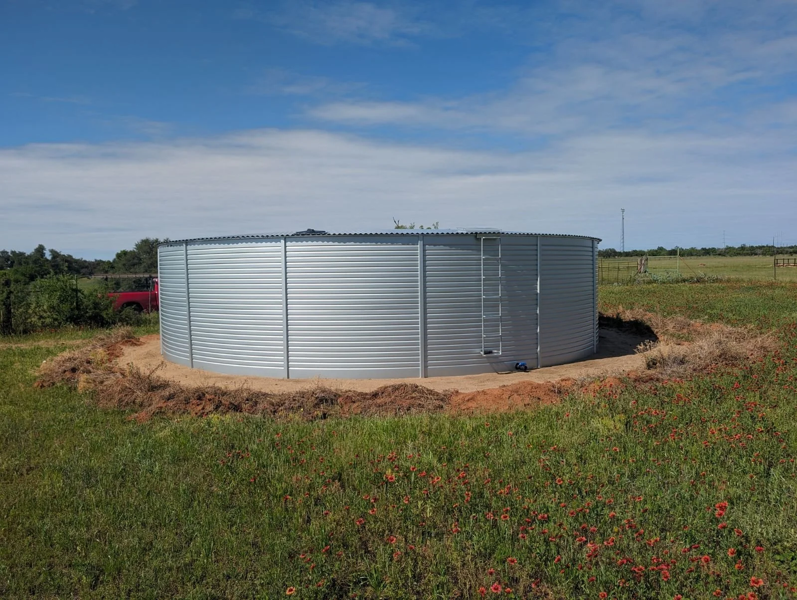 A pioneer brand rainwater storage cistern tank on a grassy field in Willow City with wildflowers, under a partly cloudy blue sky.