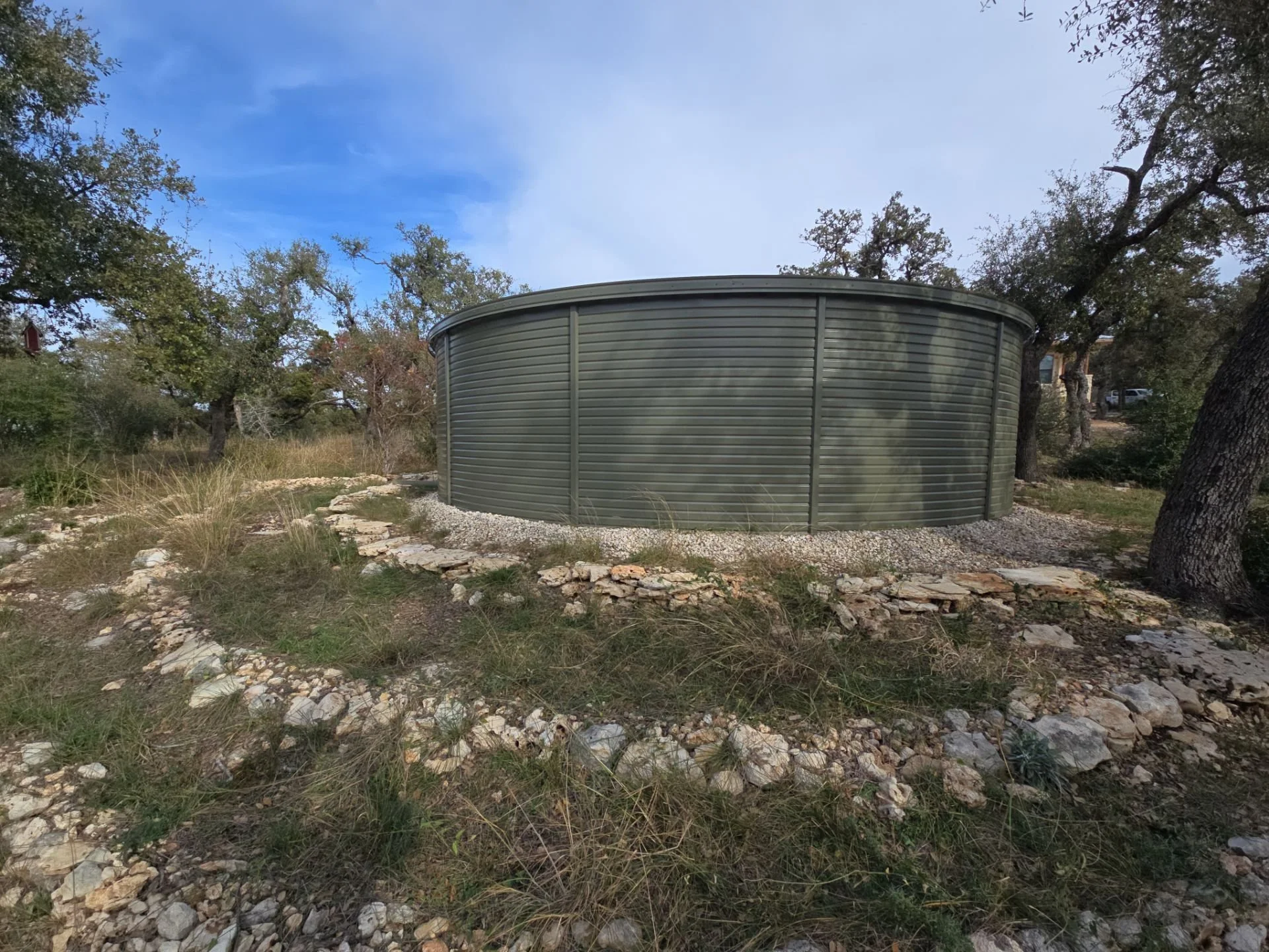 A rainwater storage cistern tank outdoors among trees and rocky terrain under a partly cloudy sky in the hill country