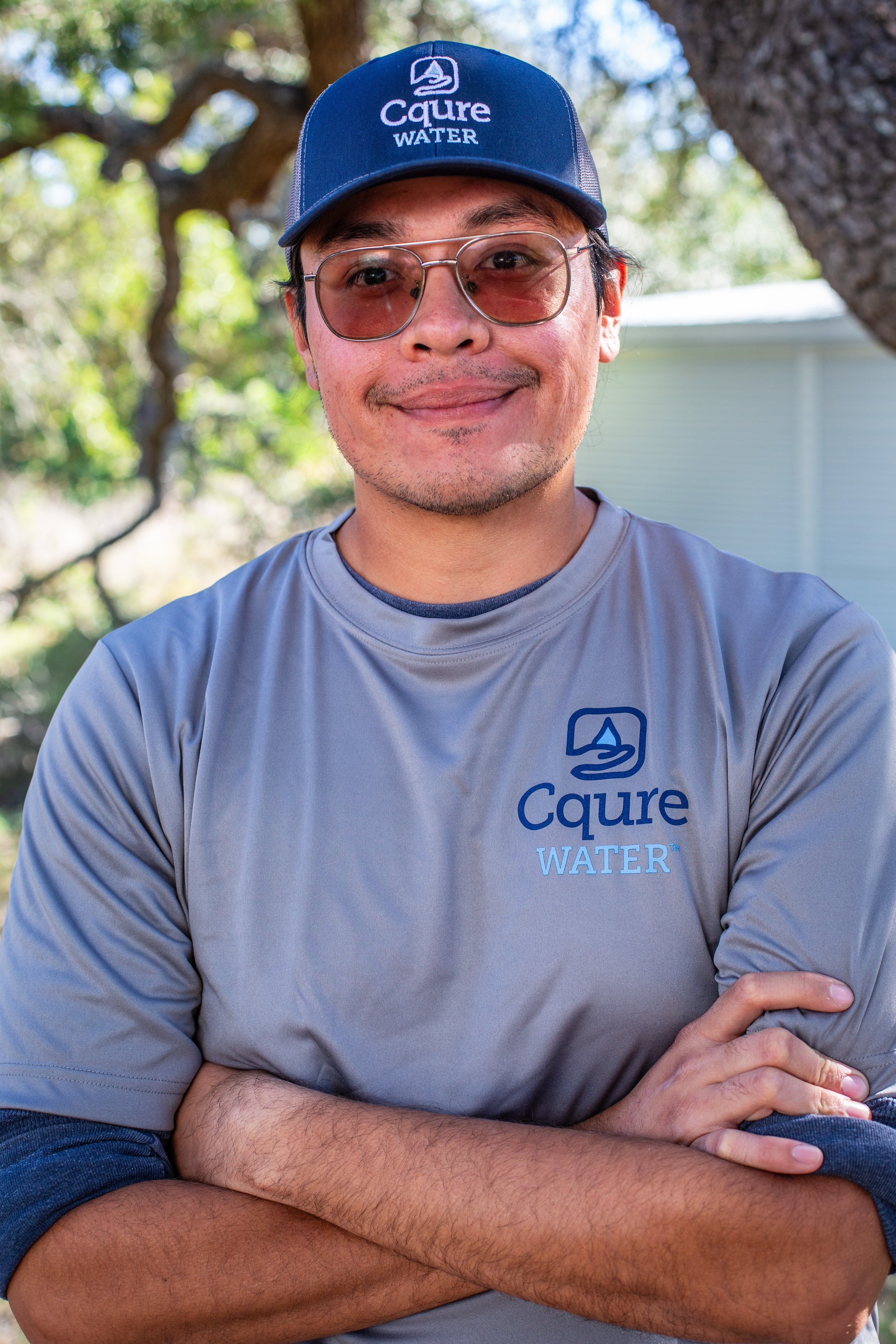 A man wearing sunglasses, a gray Caure Water t-shirt, and a blue Caure Water cap is standing outdoors with trees and a house in the background. He is smiling and has his arms crossed.