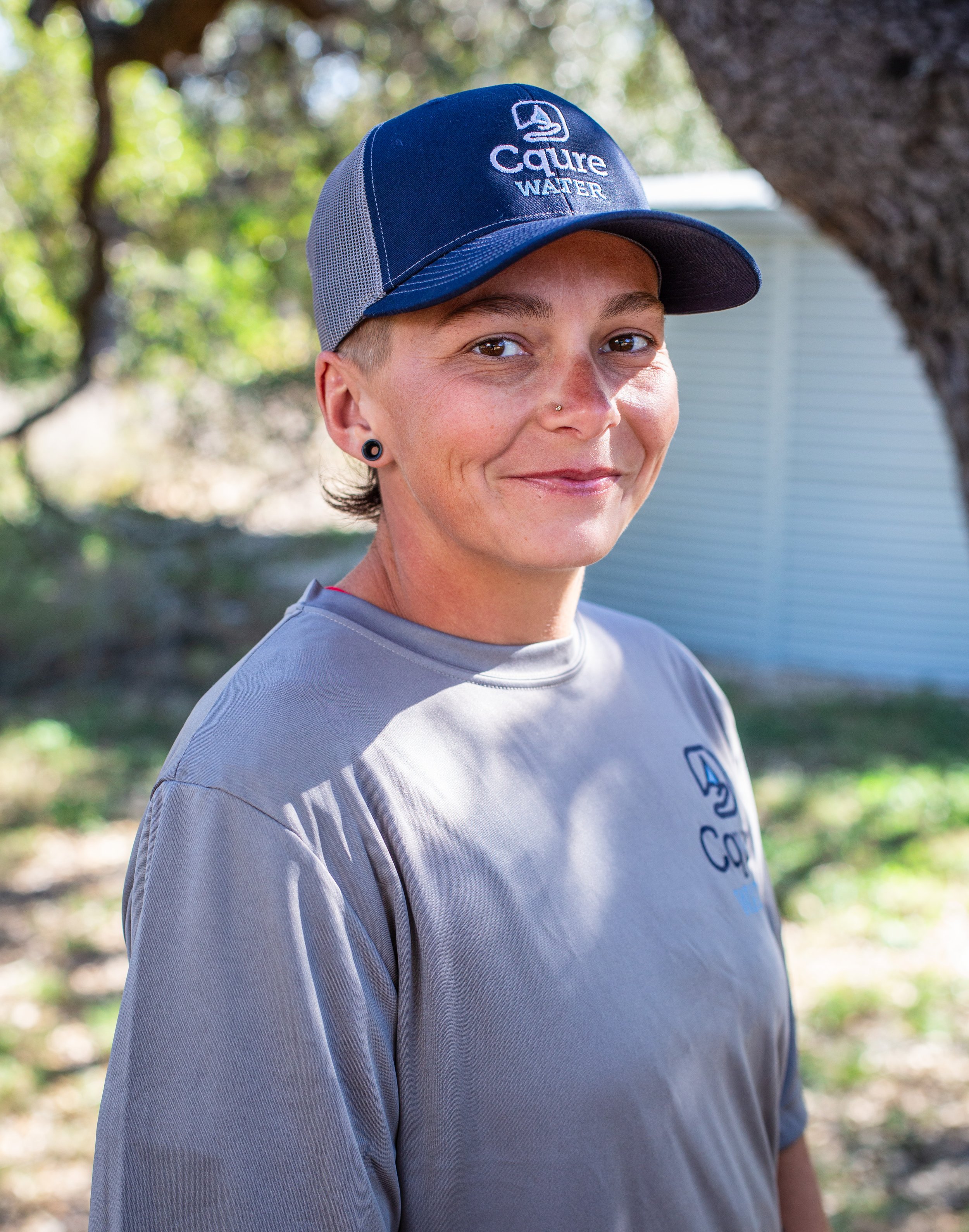 A woman with short hair and a nose piercing smiling at the camera outdoors, wearing a blue and gray cap and a gray t-shirt with a logo, standing near a tree with sunlight filtering through leaves.