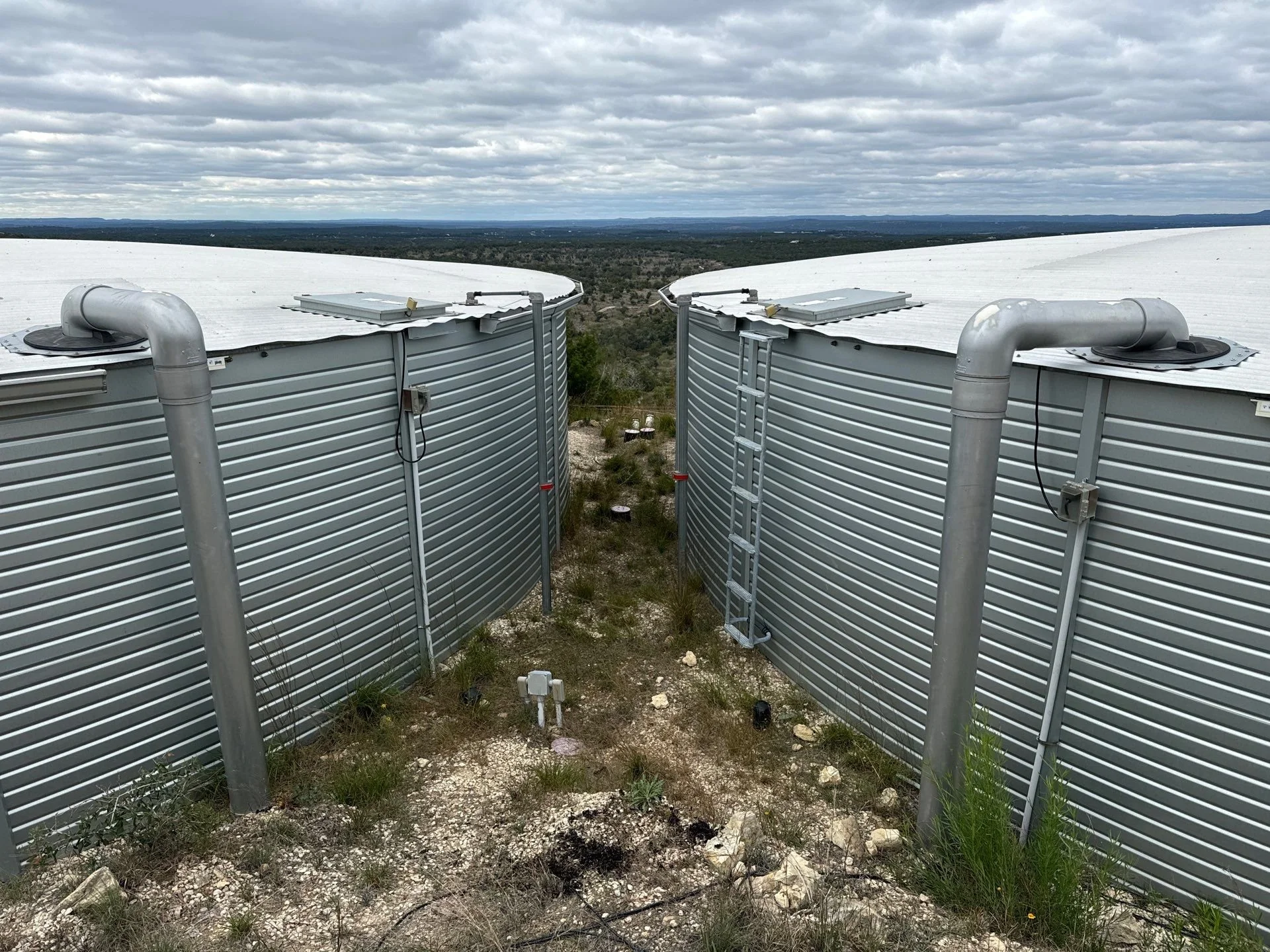 Two large pioneer brand rainwater storage cistern tanks, situated outdoors in Dripping Spring with a cloudy sky in the background.