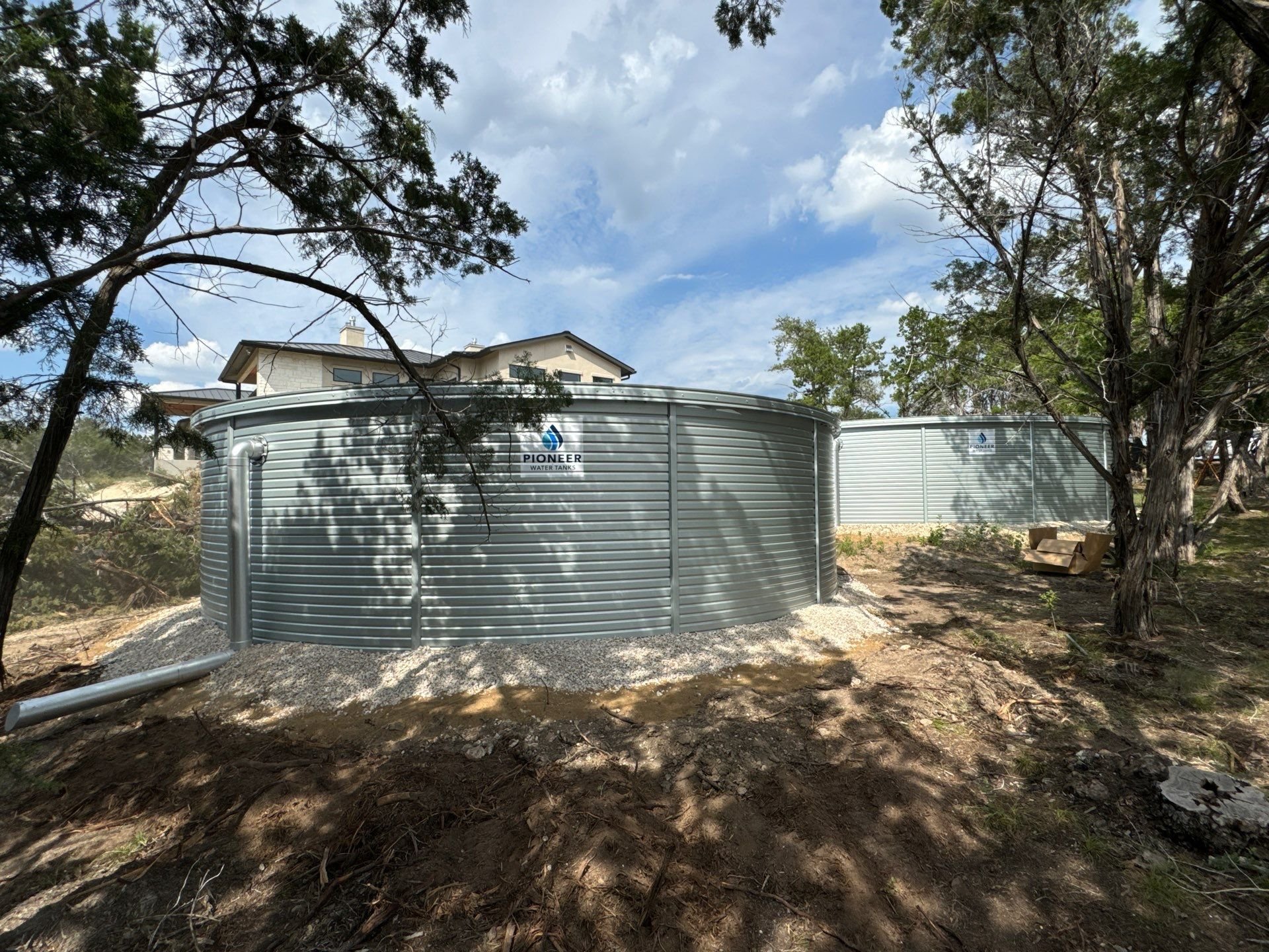 Two large Pioneer brand rainwater storage cistern tanks are situated outdoors on a slightly elevated, rocky ground surrounded by trees in dripping springs, texas. The sky is partly cloudy.