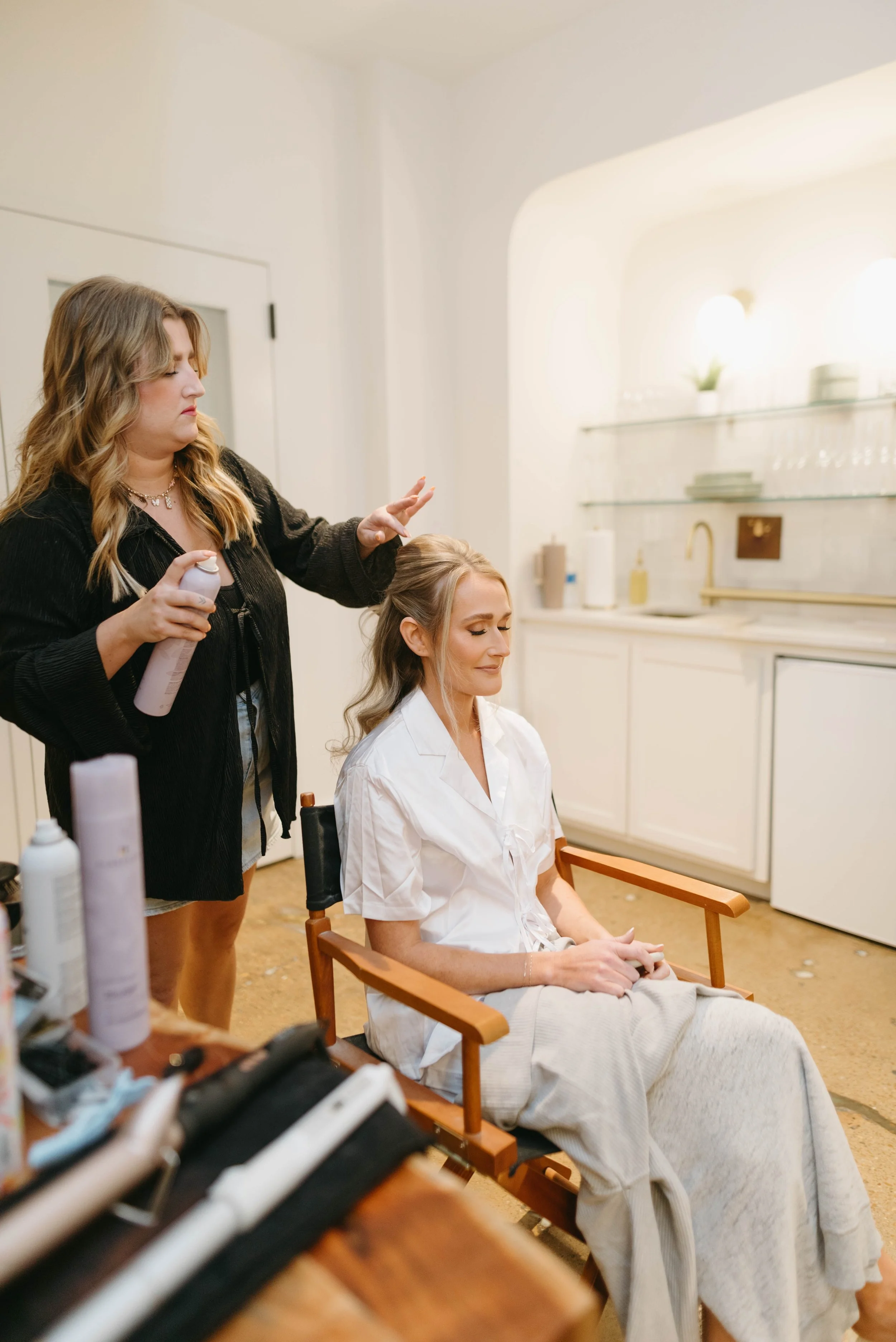 Bride in a white robe has her hair styled in a director's chair in The Suite
