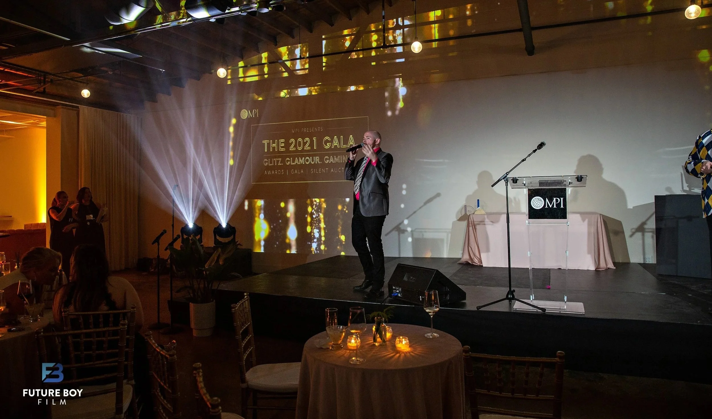 Speaker addresses a gala crowd from the stage under spotlight beams and a projected event title