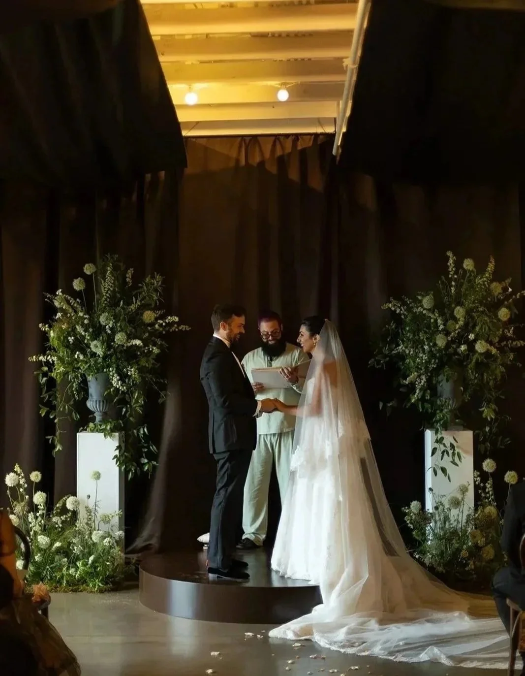 Bride and groom stand with their officiant on a round platform framed by black drapes and white floral pedestals