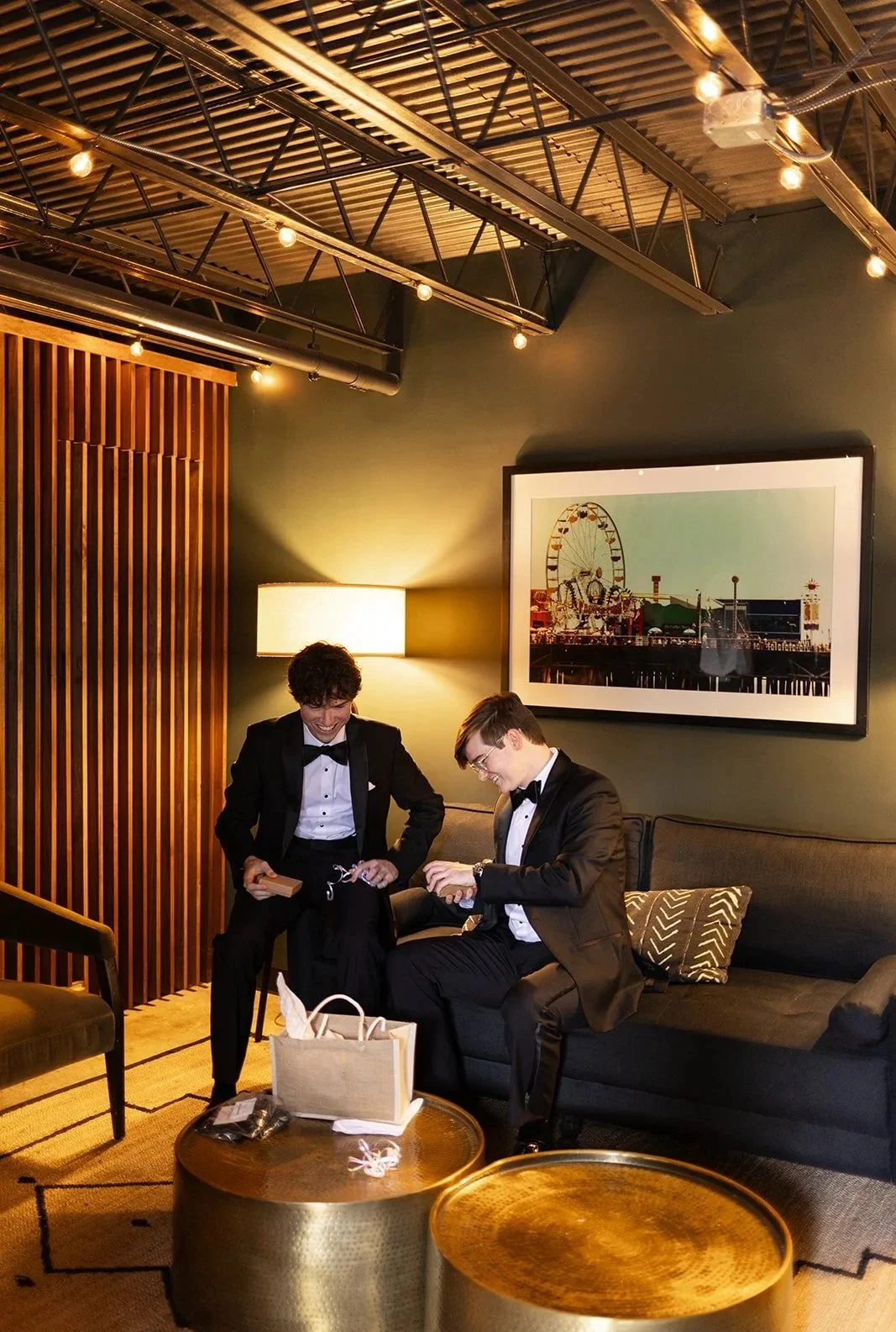 Two groomsmen in tuxedos sit on the sofa getting ready before the ceremony