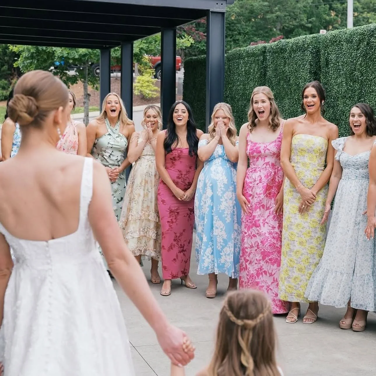 Bride in a white dress greets bridesmaids in floral dresses who react with excitement on the patio