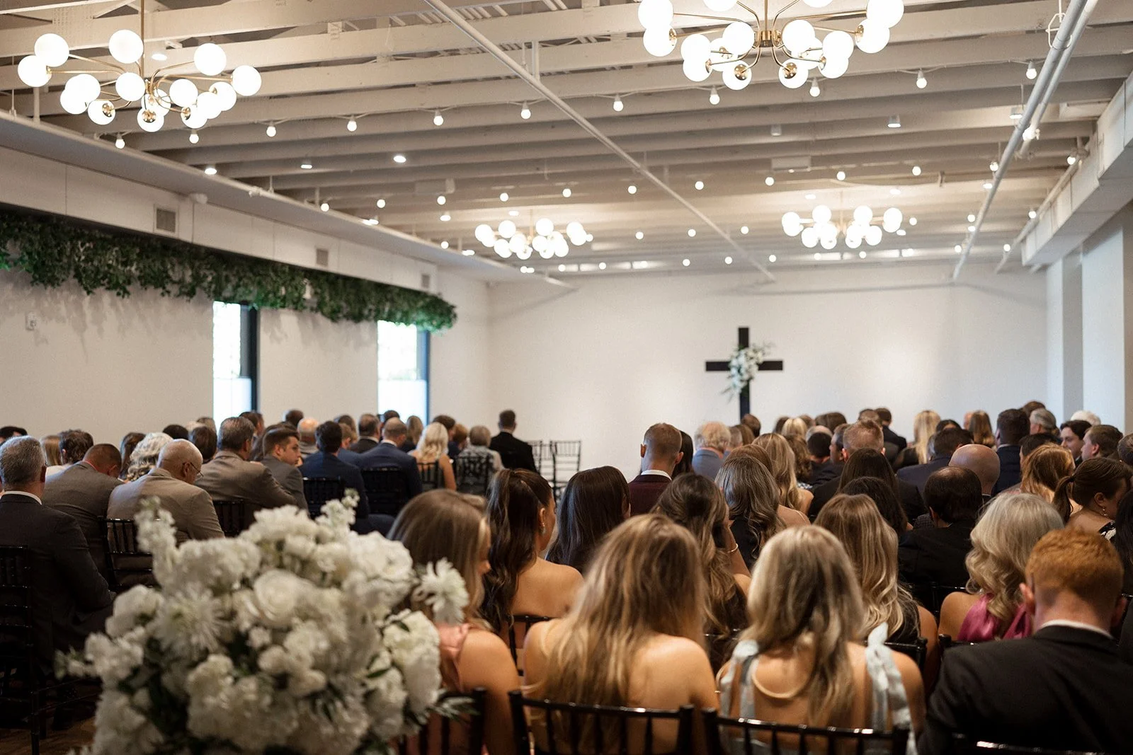 Guests seated for a wedding ceremony facing a cross on the wall beneath globe chandeliers