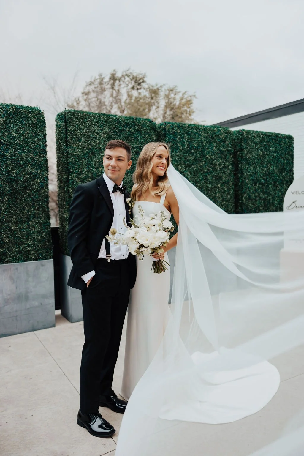Couple poses on the patio as the bride's long veil flows beside a boxwood hedge wall