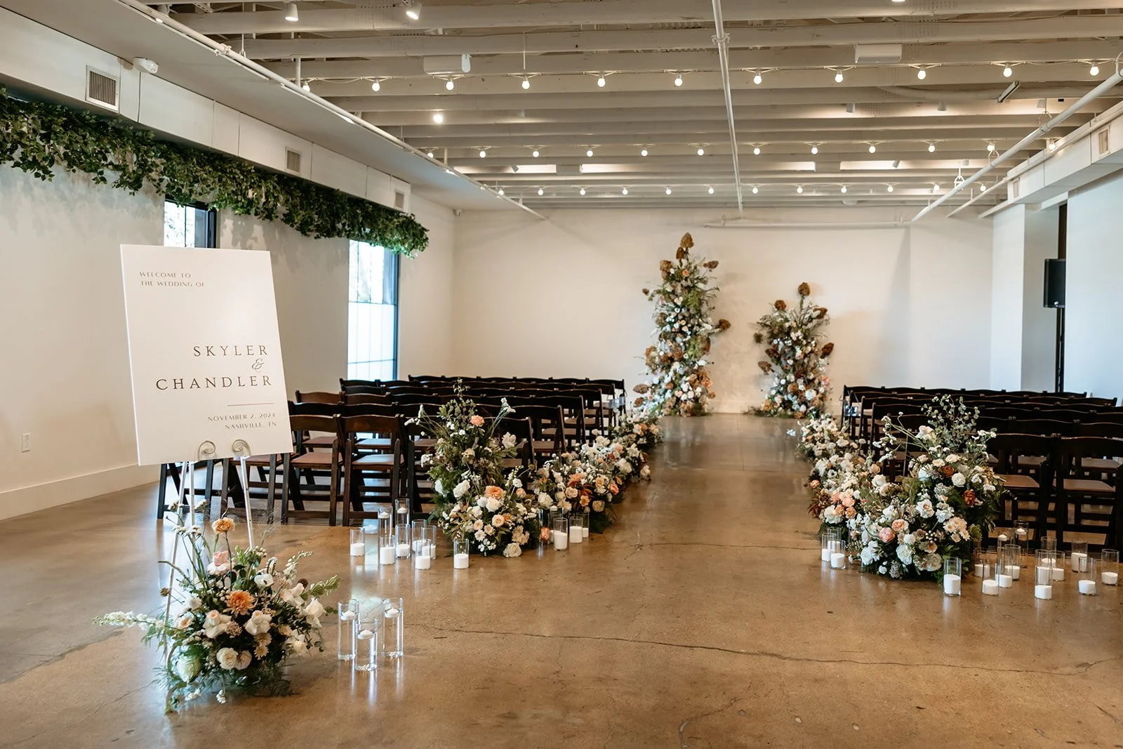 Ceremony aisle lined with candles and white floral arrangements between rows of wooden chairs, with a welcome sign on an easel