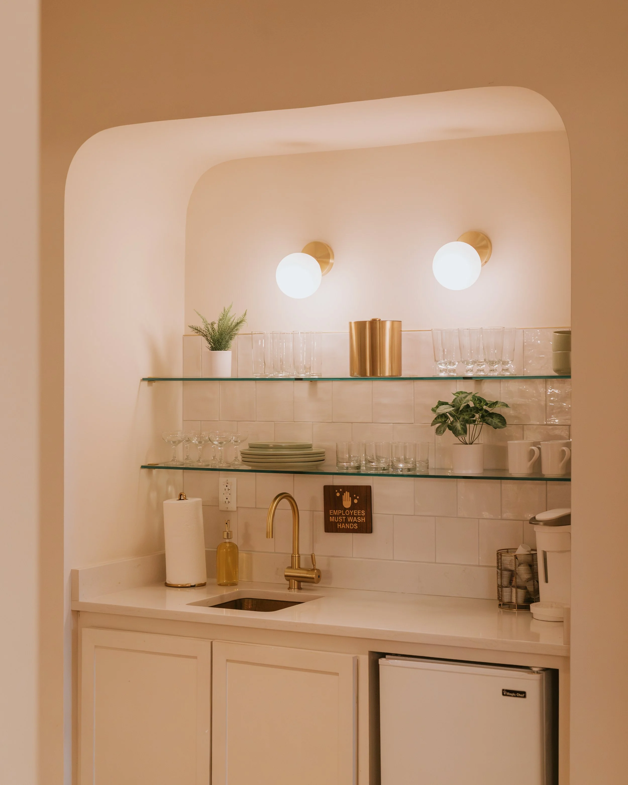 Wet bar alcove with glass shelves of glassware, brass sconces, a sink and a mini fridge