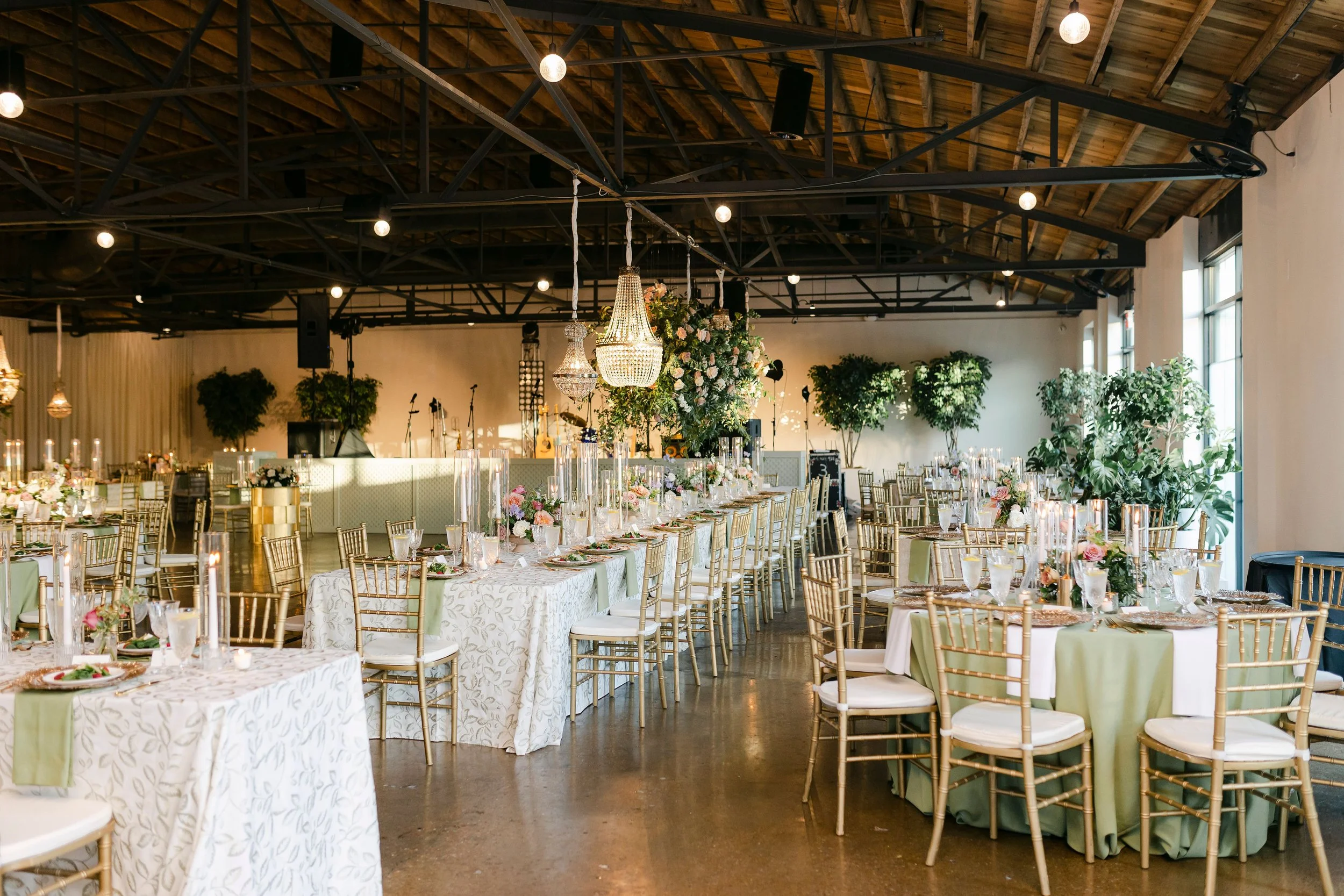 Reception tables in sage and white linens with gold chiavari chairs and greenery under the wood truss ceiling