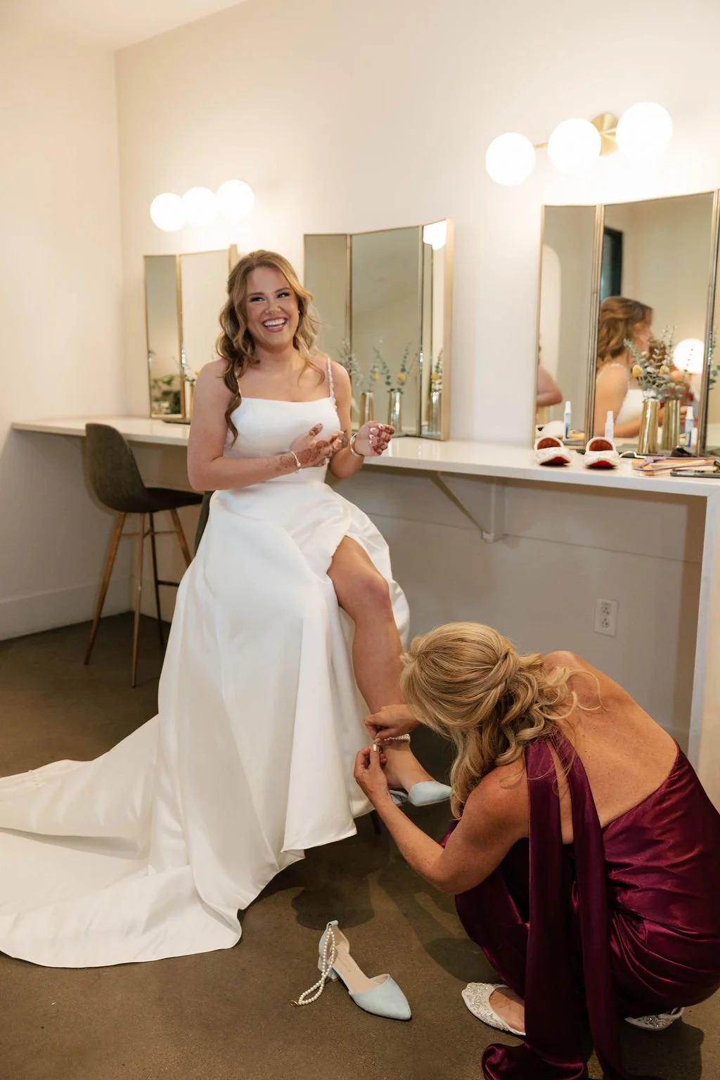 Bridesmaid in burgundy helps the bride with her shoe at a vanity with globe-lit mirrors