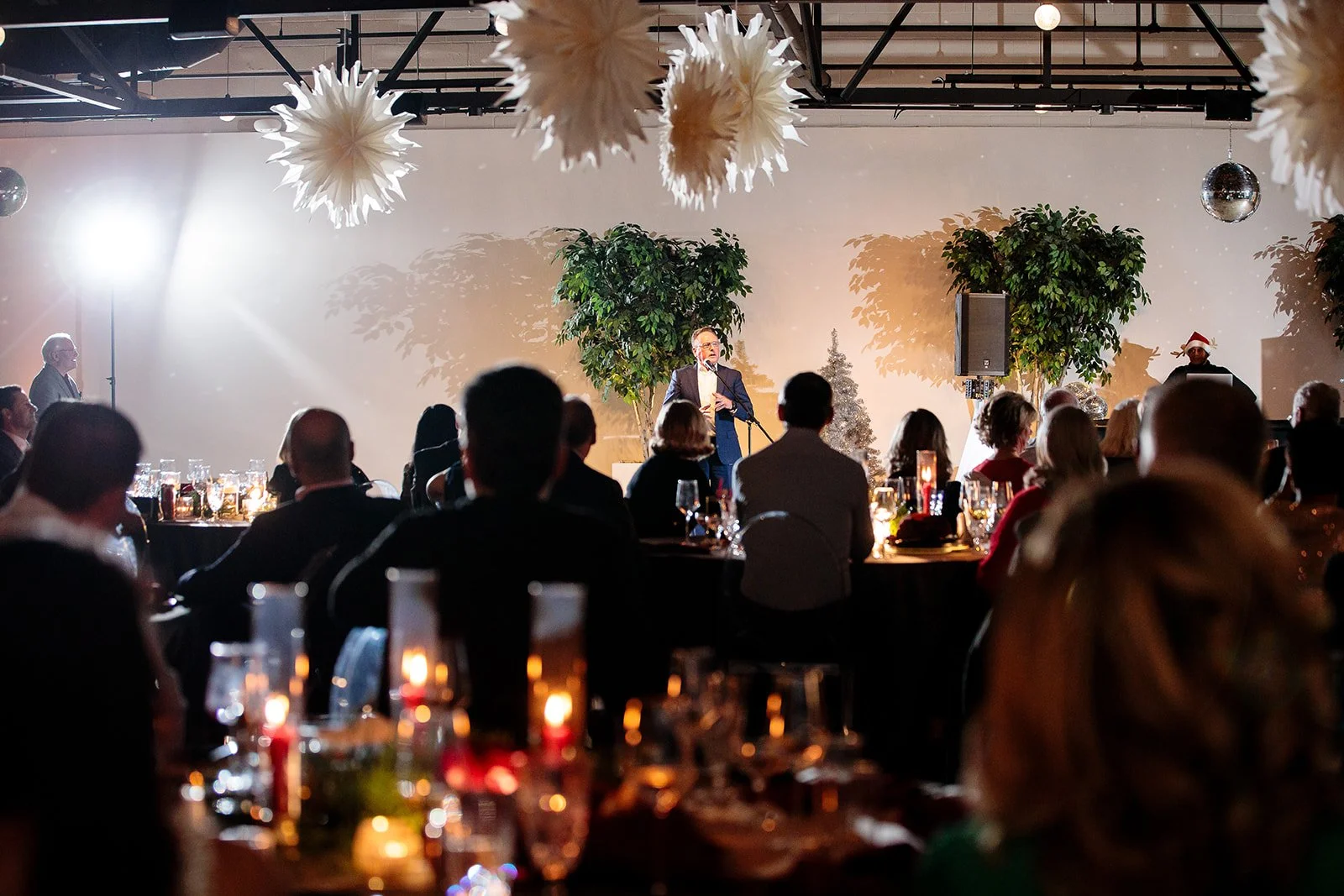 Speaker addressing guests at a holiday gala in a corporate event space with paper snowflake decor and candlelit tables