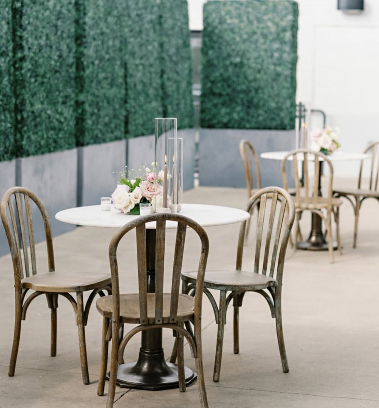 Patio cocktail tables with bentwood chairs and white flowers set against hedge walls