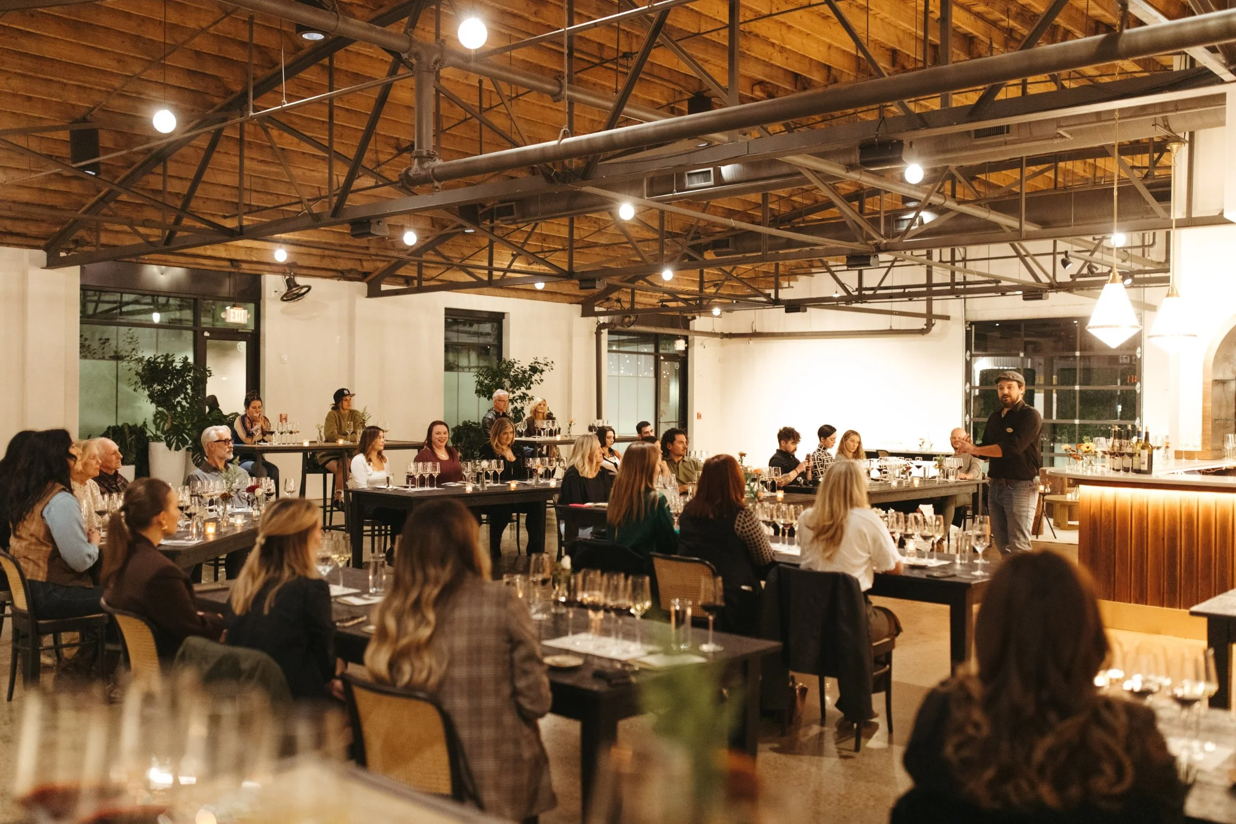 Presenter leading a seated wine tasting seminar in a corporate event space with warm lighting and industrial ceilings