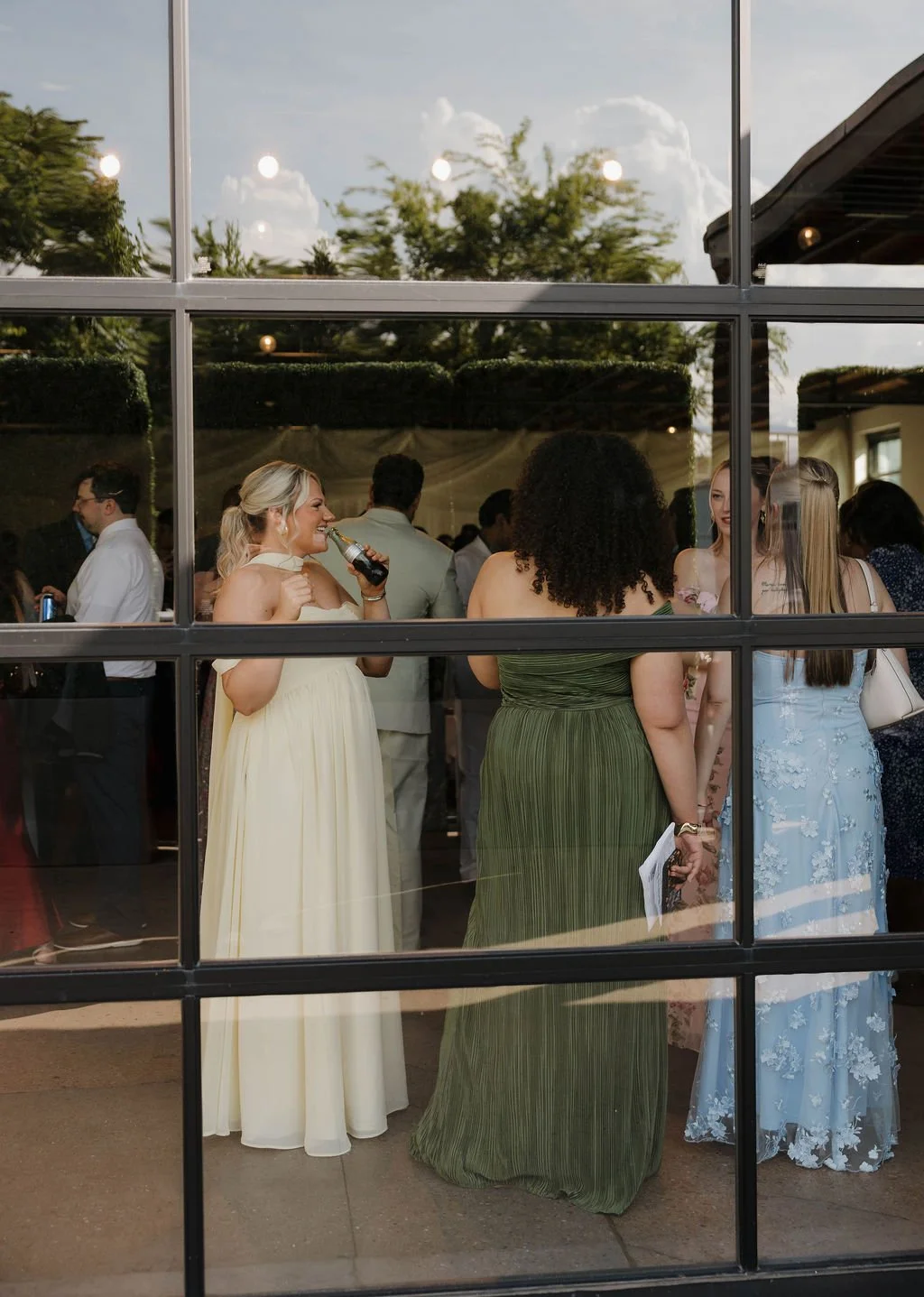 Wedding guests in formal gowns mingle, seen through Saint Elle's black-framed windows