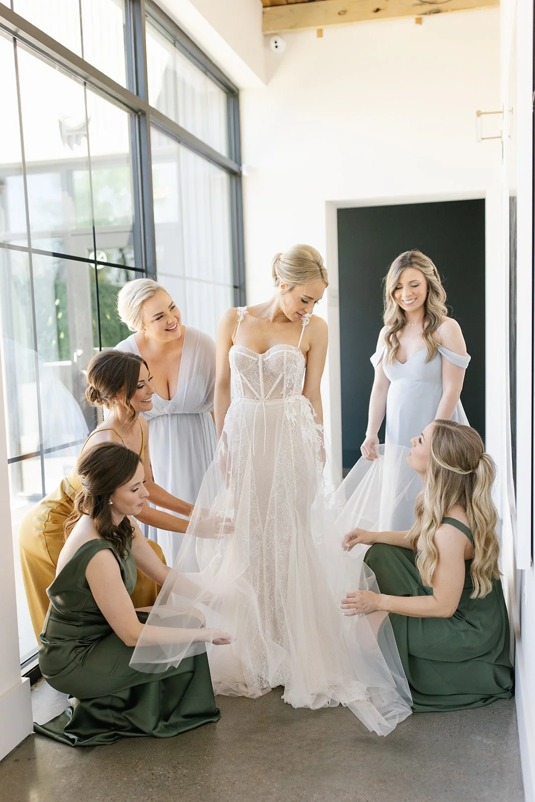 Bridesmaids in sage and blue gowns help the bride into her dress beside tall black-framed windows