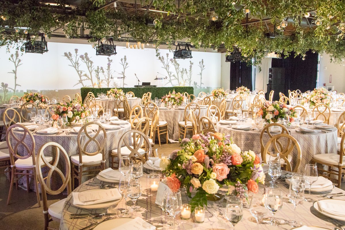 Round tables with blush floral centerpieces and gold cross-back chairs under a greenery canopy