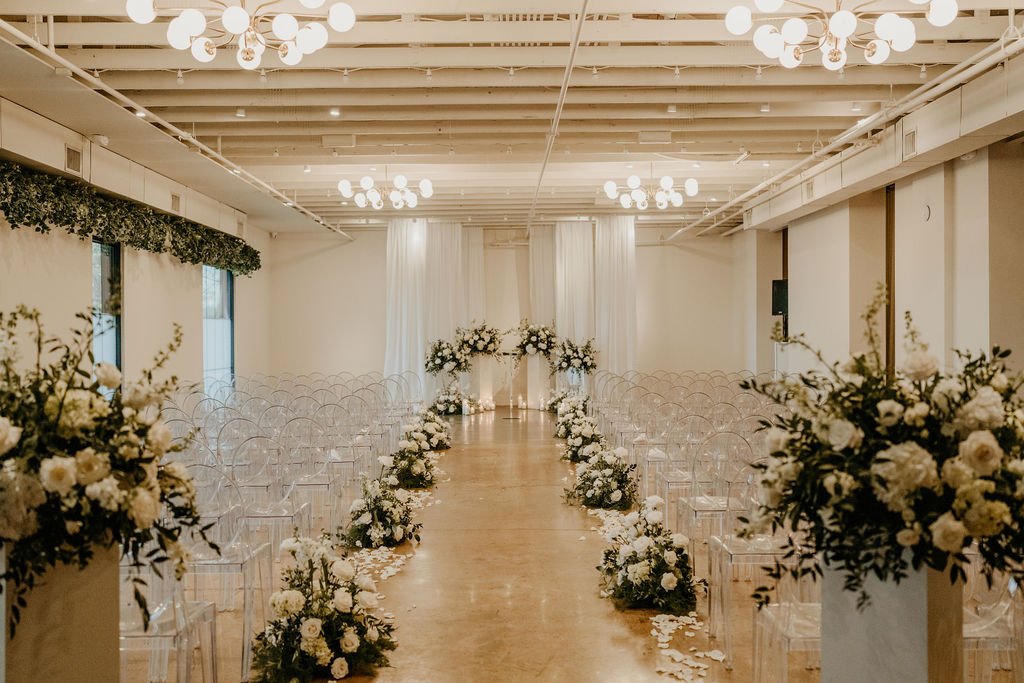 White flower arrangements and clear chairs frame a ceremony aisle in Saint Elle's light-filled Conservatory