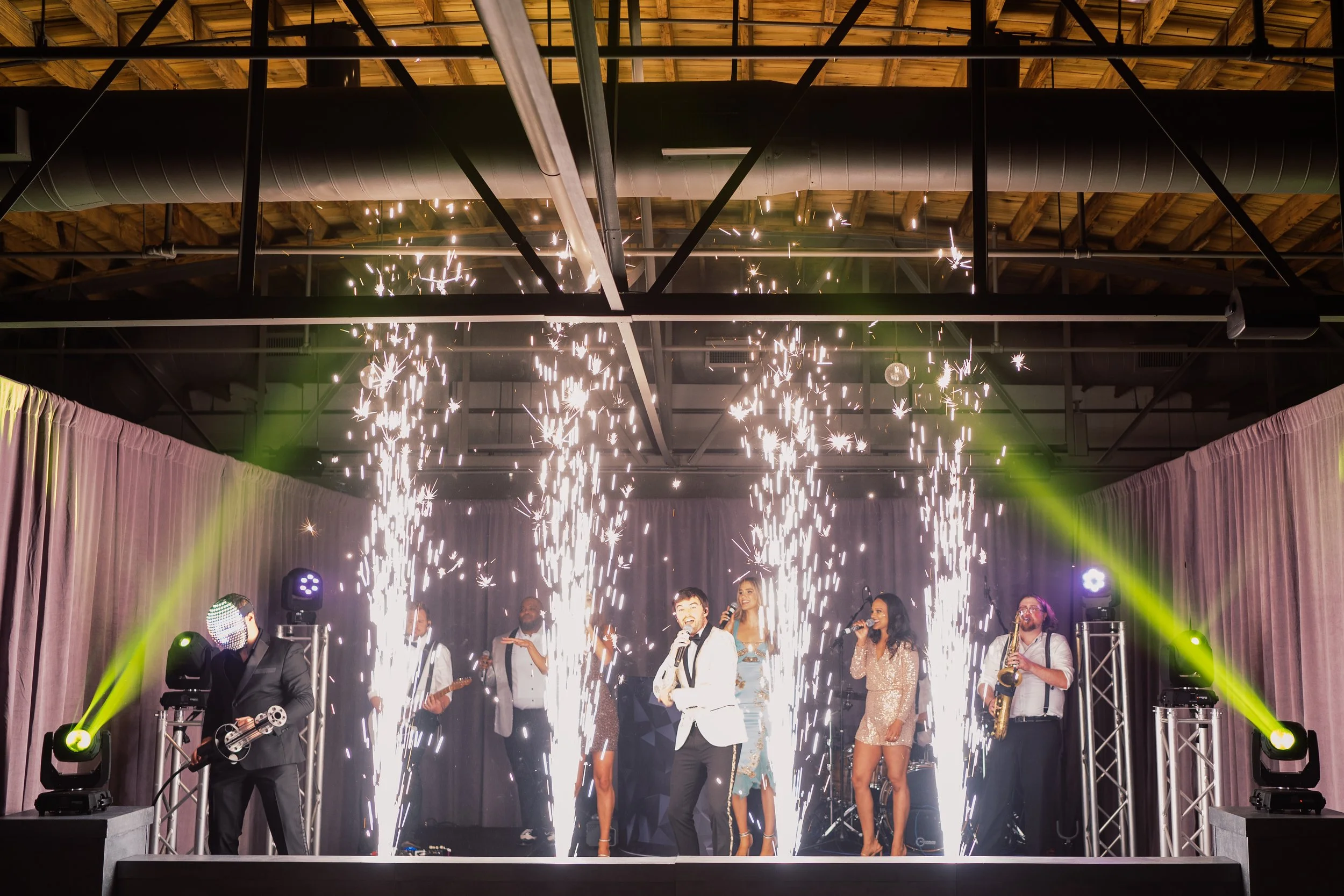 Live band performs under cold spark fountains and green stage lights against pink drapes