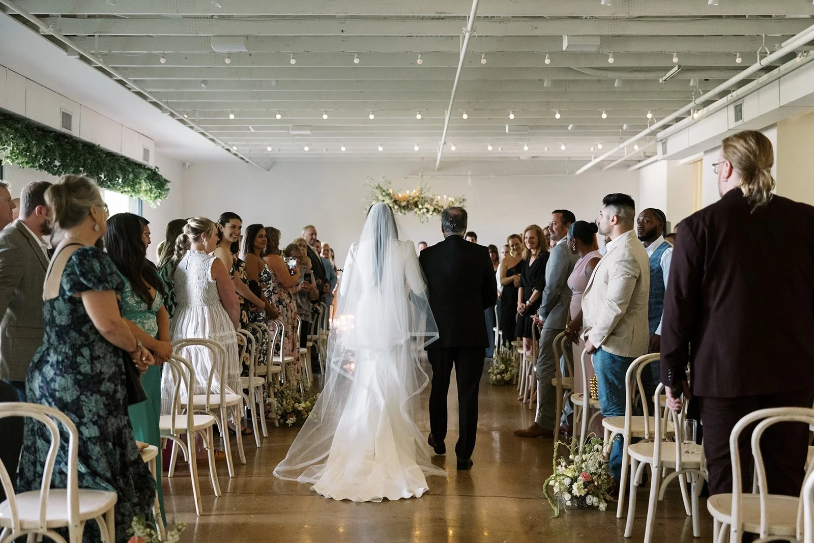 Bride in a long veil walks down the aisle on the arm of a man in a dark suit as guests stand