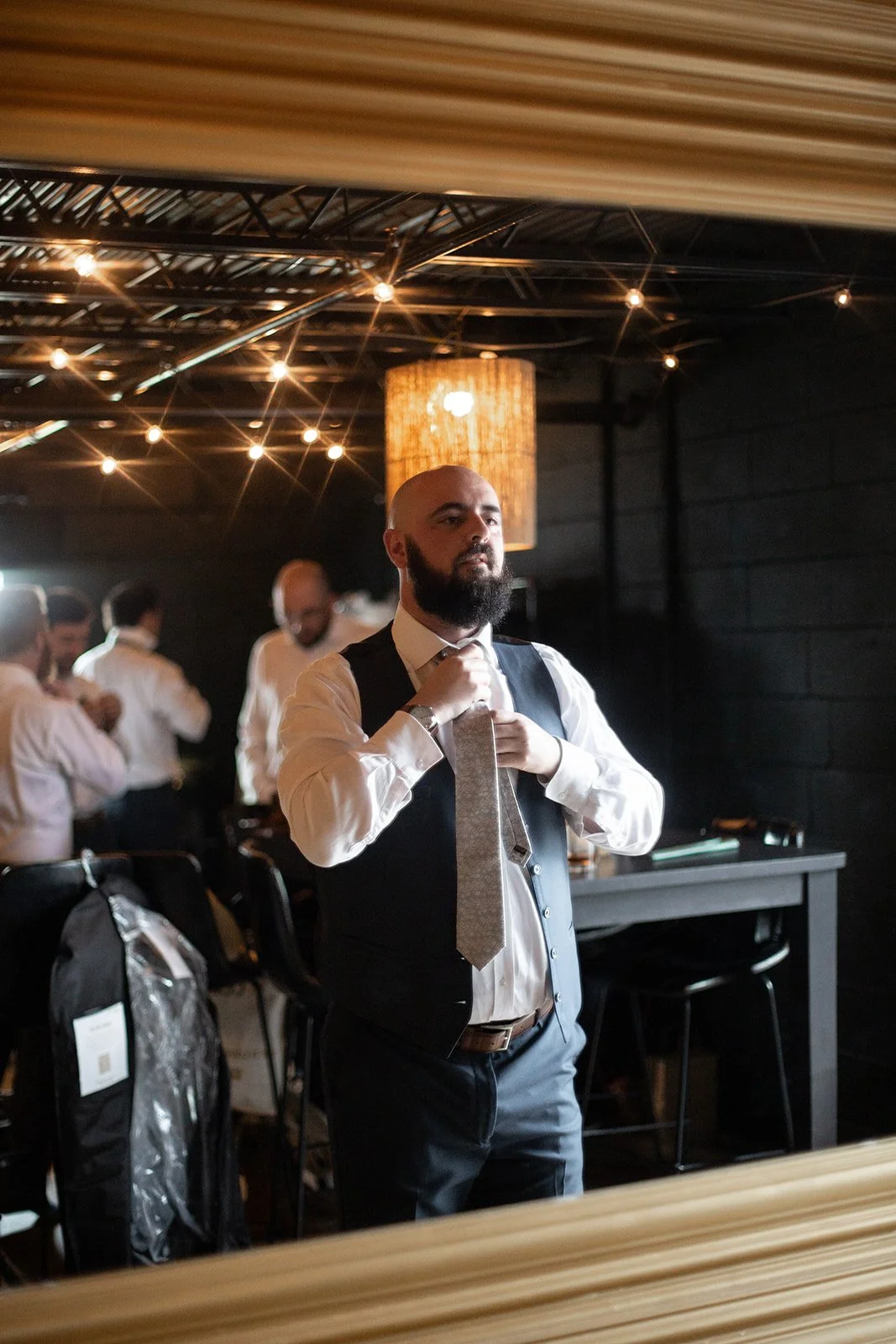 Groomsman adjusts his tie in the mirror while the wedding party gets dressed behind him