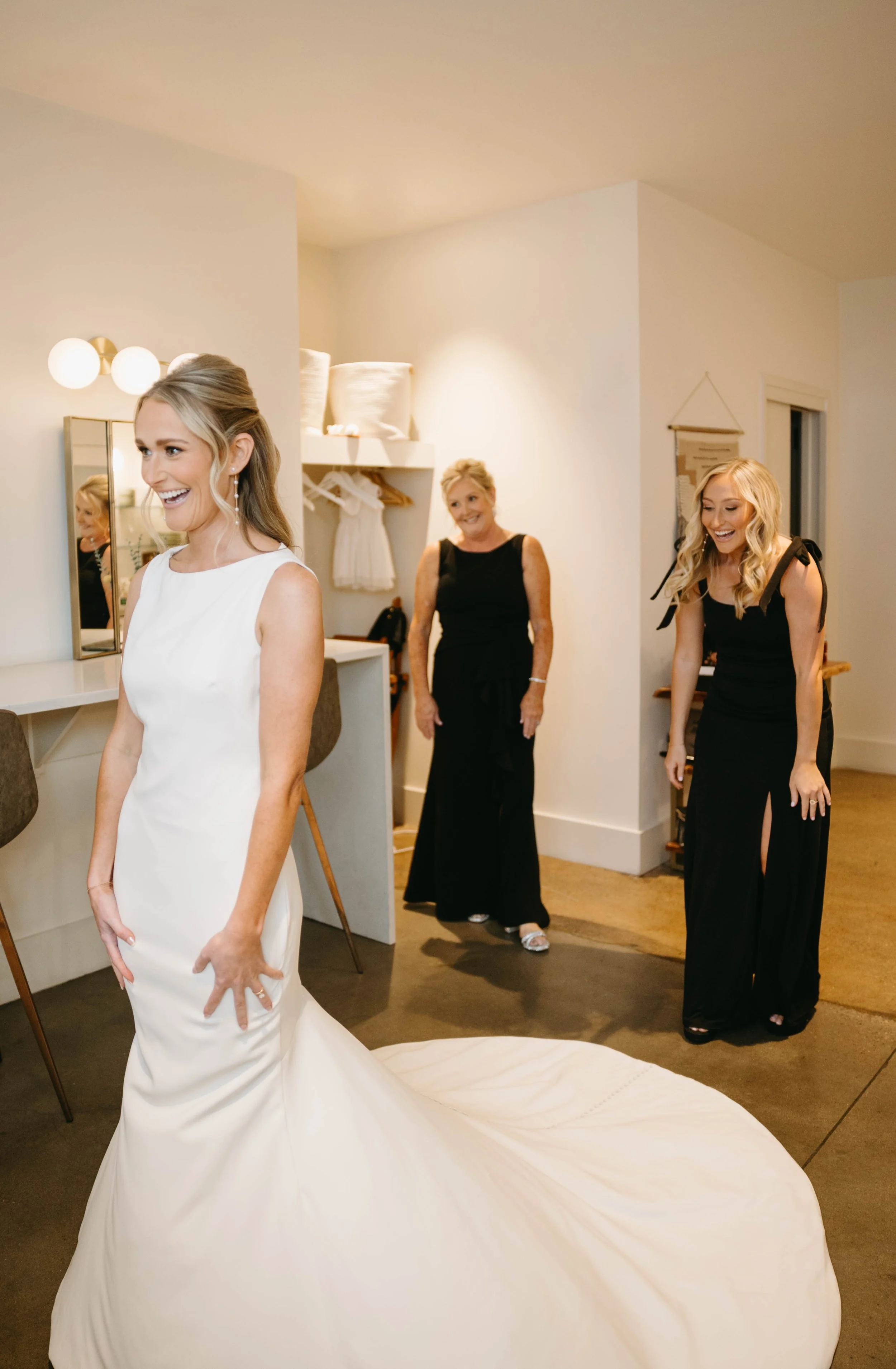 Bride in a fitted gown with a long train stands as bridesmaids in black look on