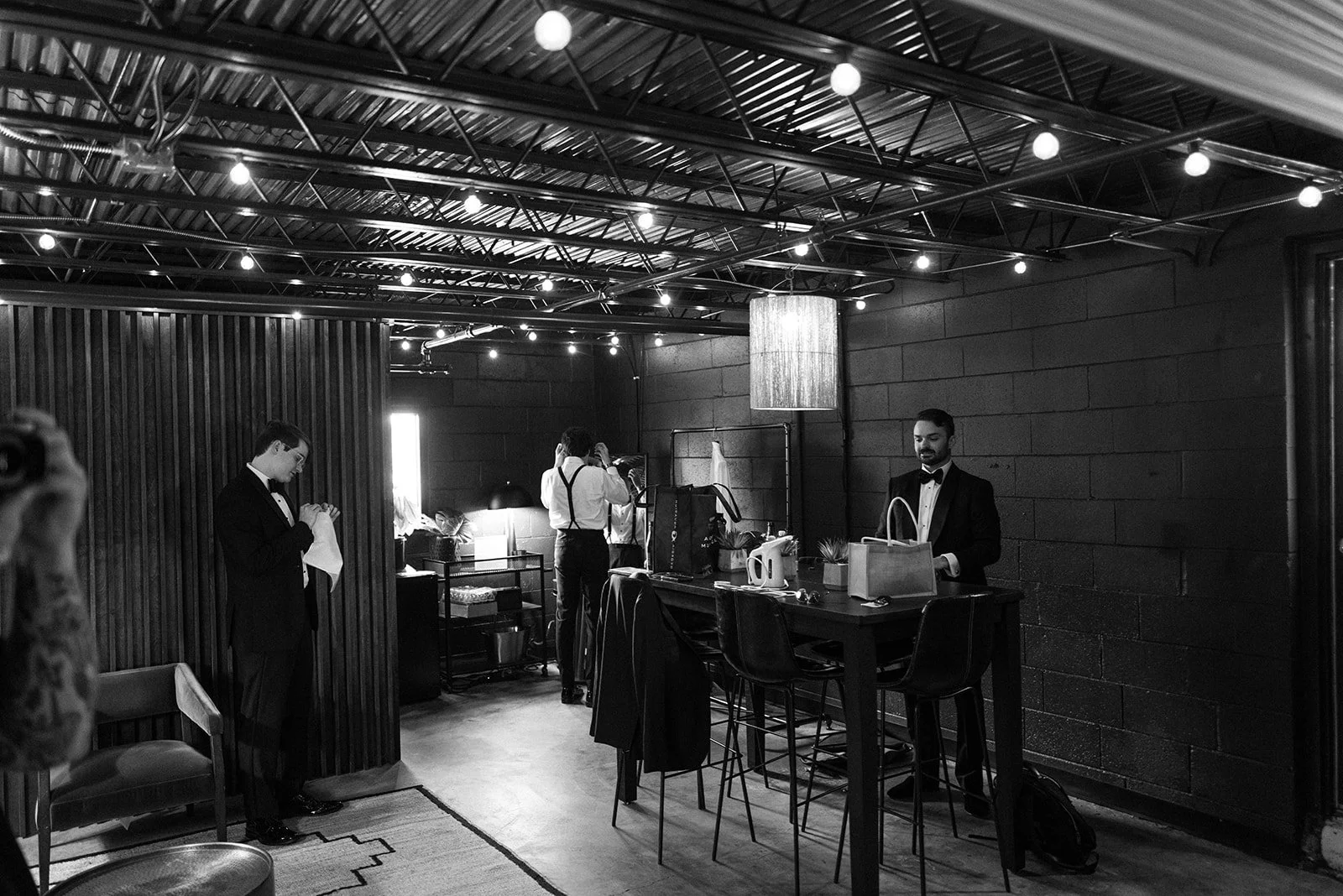 Black-and-white photo of groomsmen getting ready around a pub table under string lights