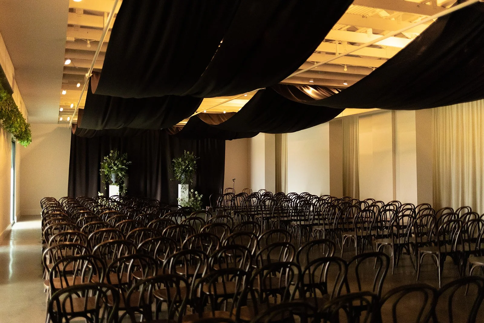Rows of black bentwood chairs face a black draped backdrop with fabric swags overhead for a moody evening ceremony