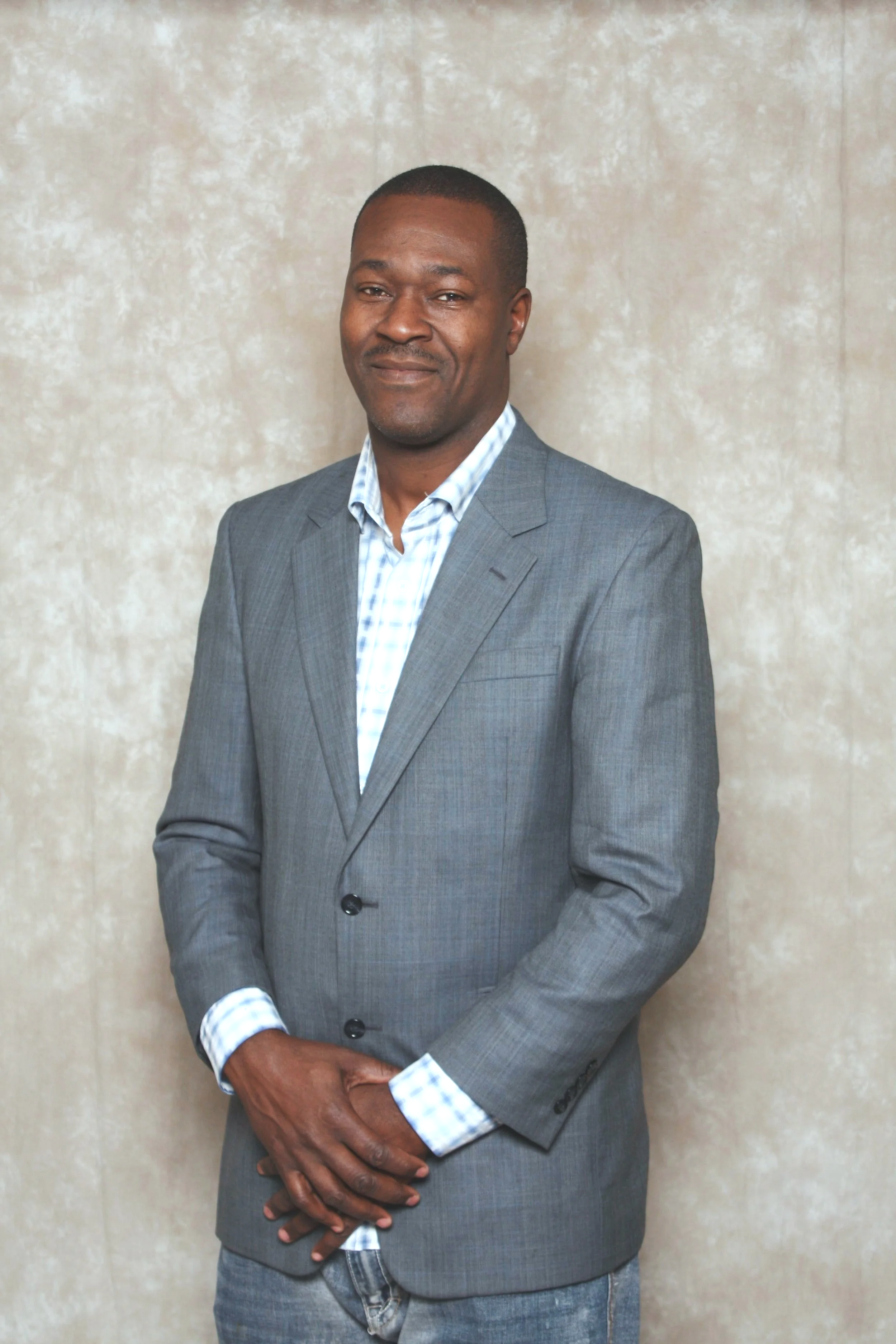 A man in a gray suit with a blue checkered shirt standing against a neutral beige background.