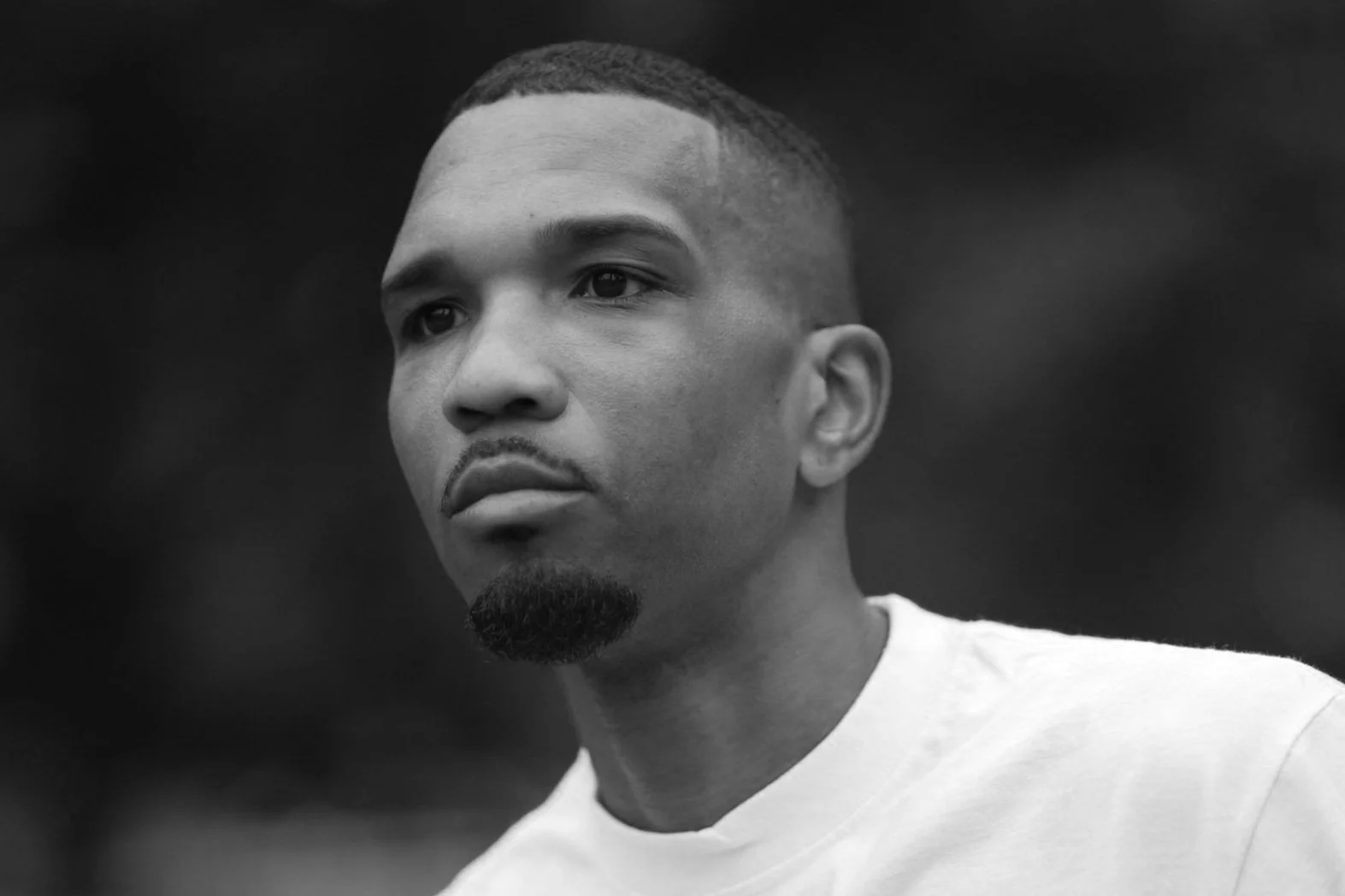 Black and white portrait of a young man with short hair, a goatee, wearing a white shirt, looking pensively into the distance coming from a pickleball game in NYC.