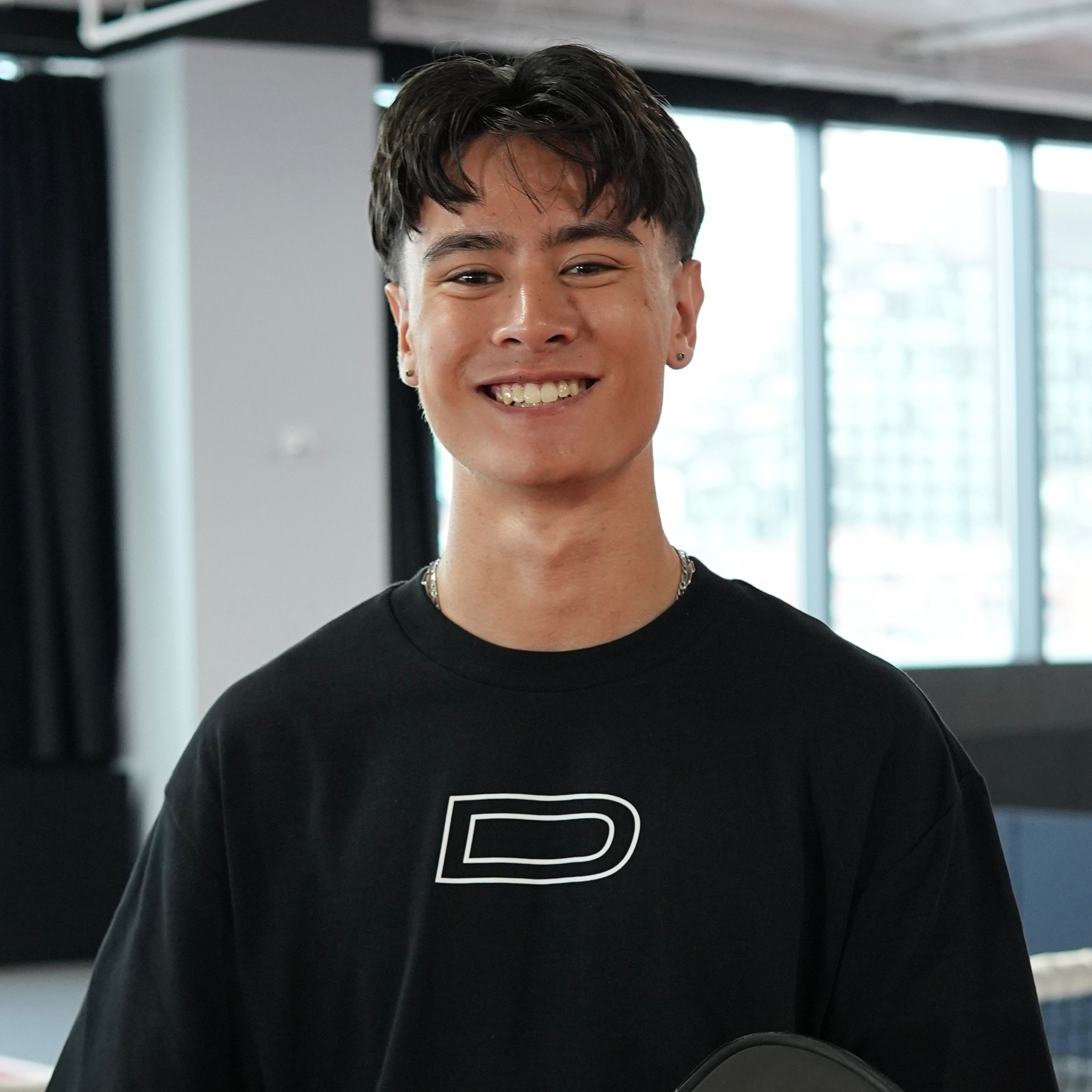 Black and white portrait of a young man with short hair, a goatee, wearing a white shirt, looking pensively into the distance coming from a pickleball game in NYC.
