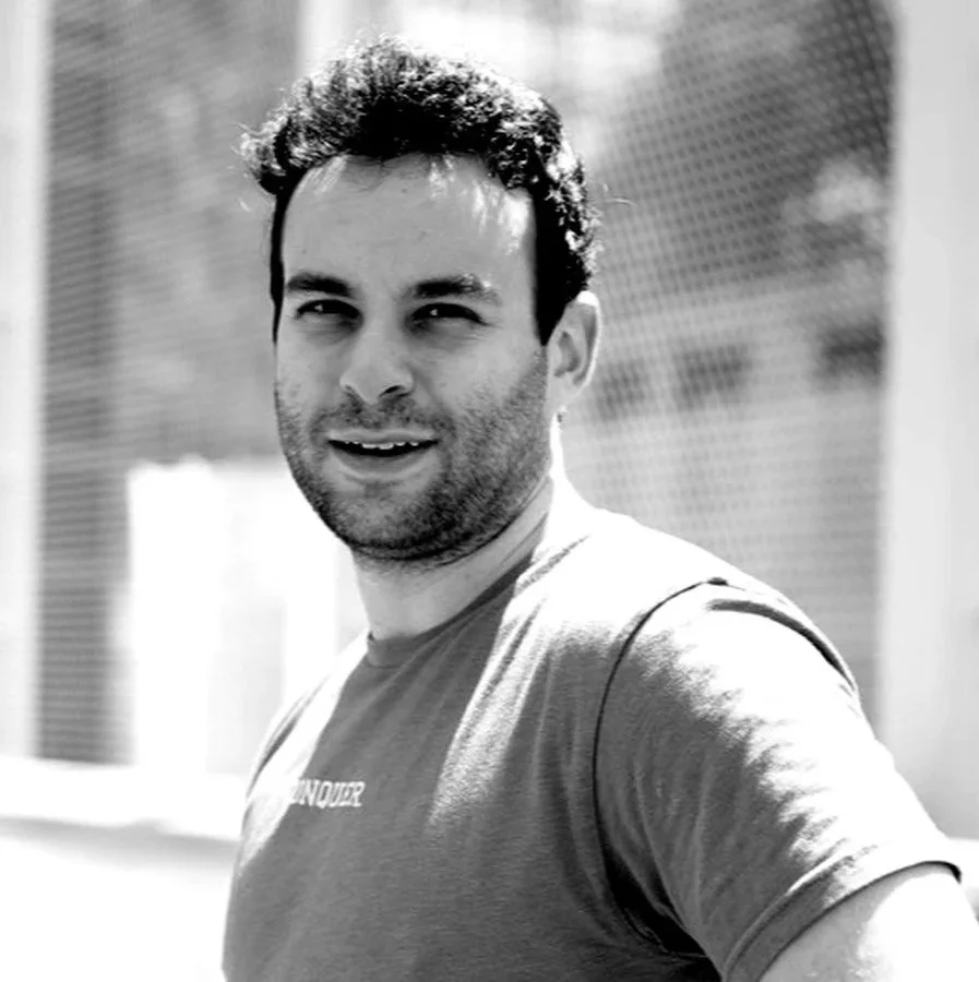 A black and white photo of a man wearing sunglasses, standing in front of a chain link fence playing pickleball.