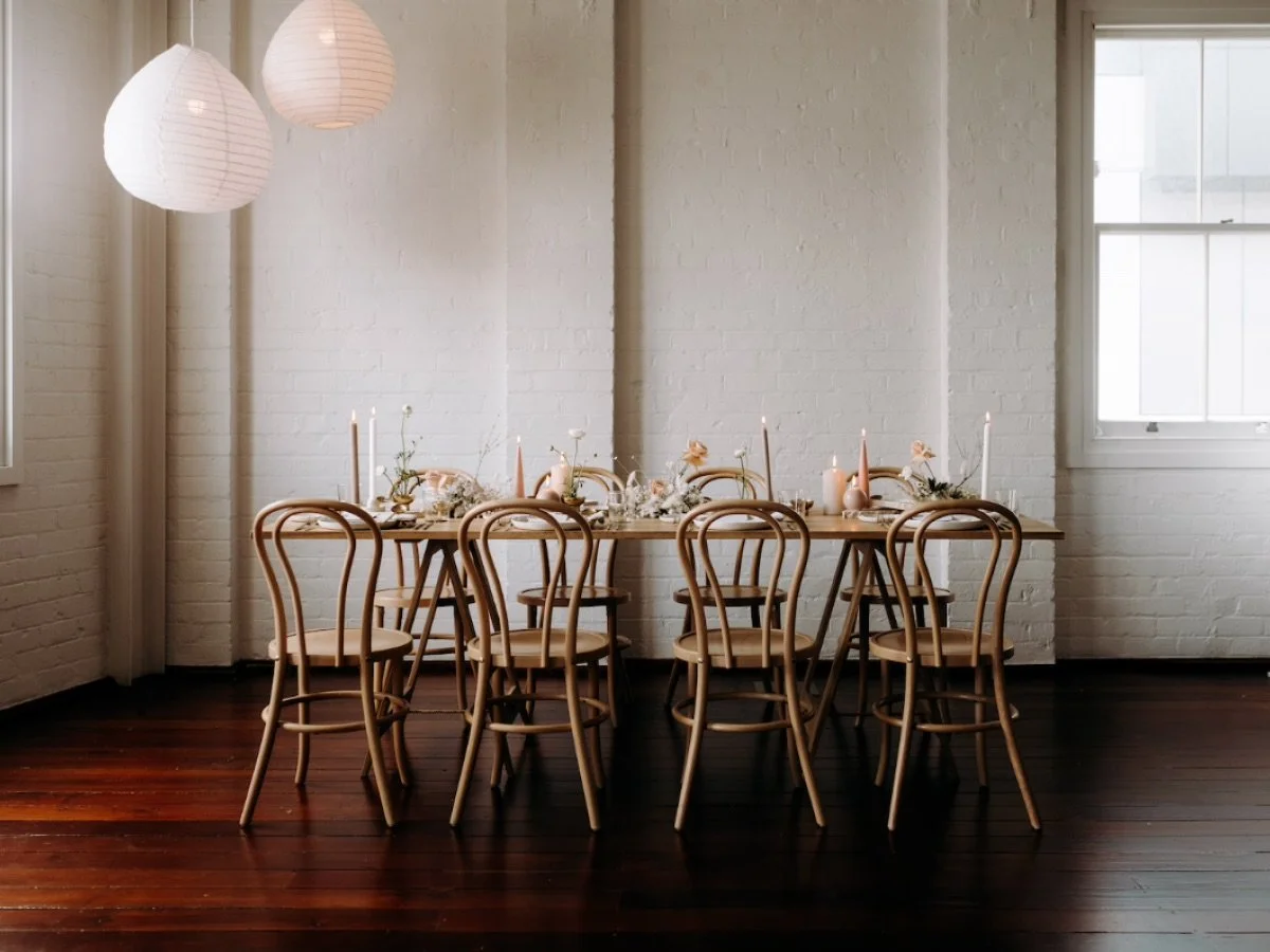Minimalist dining room with a wooden table, six chairs, white brick walls, large windows, and hanging paper lanterns. The table is set with candles and floral arrangements.